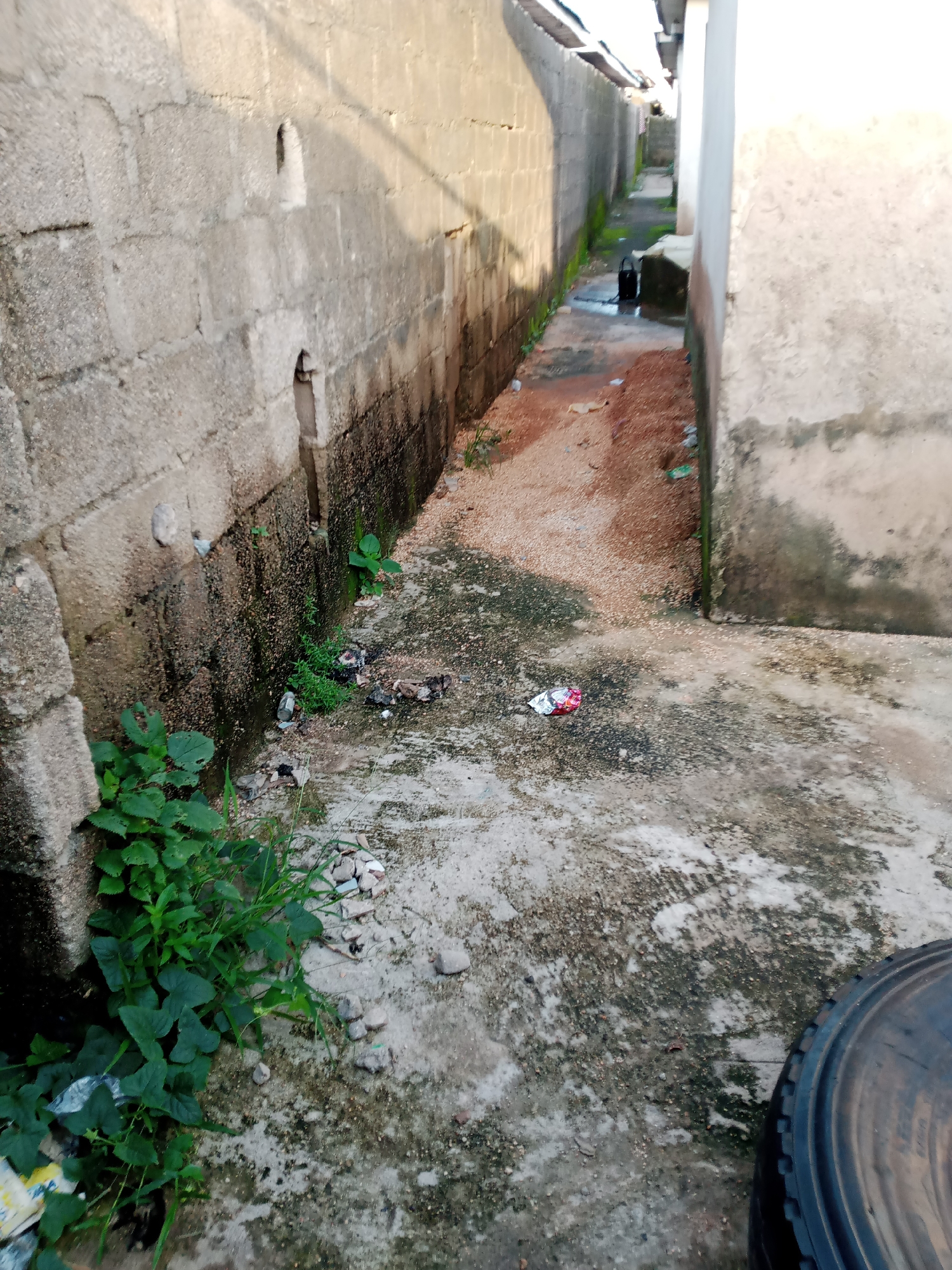 The image depicts a narrow alleyway in Jauro Yinu, Nigeria. The alley is flanked by concrete block walls on both sides. The ground is a mixture of concrete and loose gravel, with some patches of dirt and vegetation. Scattered debris, including plastic wrappers and what appears to be trash, litters the alley. In the distance, a black bucket is visible on the ground. A portion of a black tire is visible in the foreground, on the right side of the image. The scene suggests an outdoor, possibly residential or commercial, area within the city.