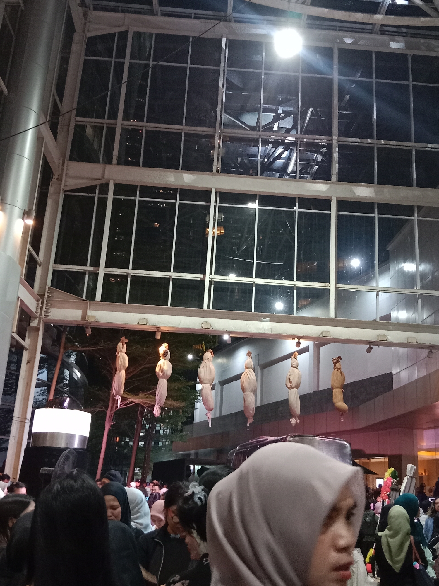 The image captures an outdoor evening scene in South Jakarta, Indonesia. A crowd of people, many wearing headscarves, are gathered. In the background, there is a large building with extensive glass windows. Hanging from a structure above the crowd are several figures wrapped in light-colored fabric, resembling effigies. The lighting suggests artificial illumination from lamps and building lights. The environment appears to be an urban public space, possibly a plaza or walkway between buildings, with trees visible in the background.