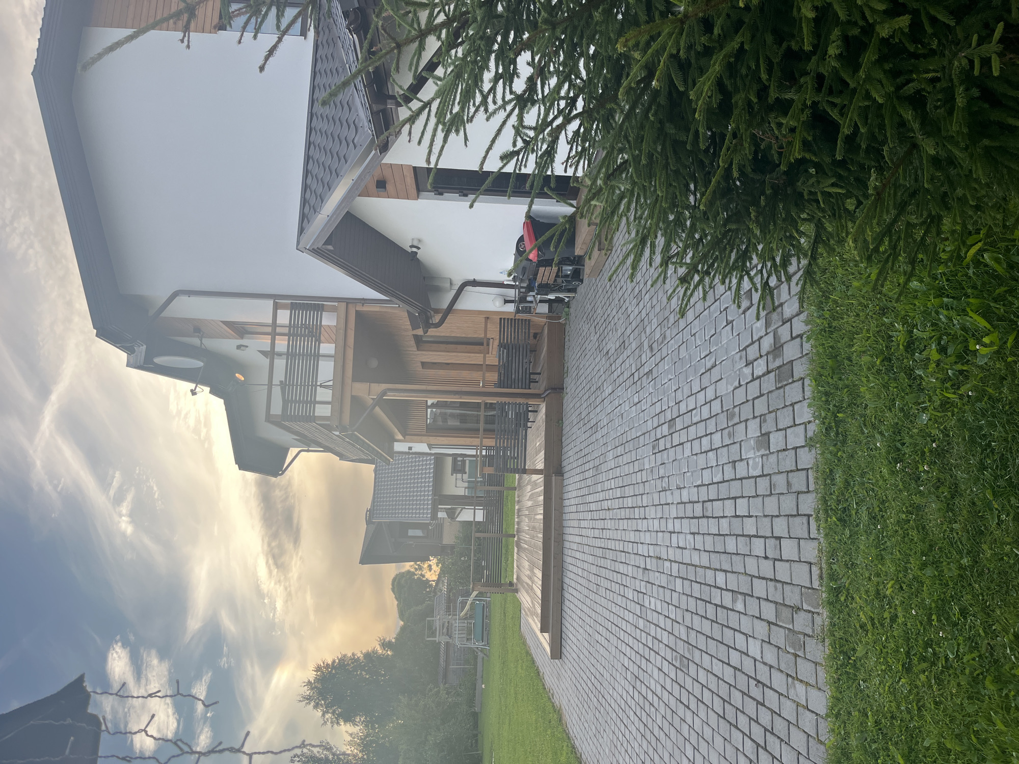 The image depicts the exterior of a modern, multi-story house in Vladimirovka, Russia. The house features white siding with wooden accents and dark roofing. In the foreground, a paved driveway made of grey interlocking pavers extends towards the house. To the left of the driveway is a neatly mowed green lawn. Adjacent to the house is a wooden deck with railings. Further in the background, more houses and trees are visible under a sky with scattered clouds and a hint of sunset. A dark green fir tree is prominent in the right foreground. Some outdoor equipment, including a grill and trash cans, are situated near the side entrance of the house. There are no people visible in the image. The overall scene suggests a residential property in a suburban or semi-rural setting.