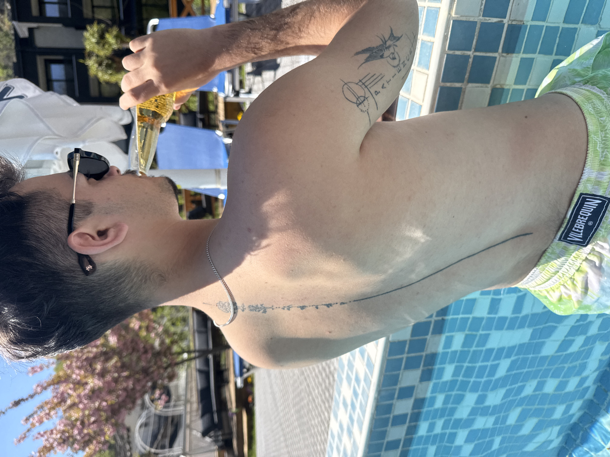 A person is seen drinking from a bottle while sitting by a blue-tiled pool. The individual is wearing sunglasses and has a tattoo on their left arm. The person is wearing green swimming trunks with the brand name "VILEBREQUIN" visible. The background shows trees and buildings, possibly in Marchevo, Bulgaria. The picture is taken outdoors in daylight.