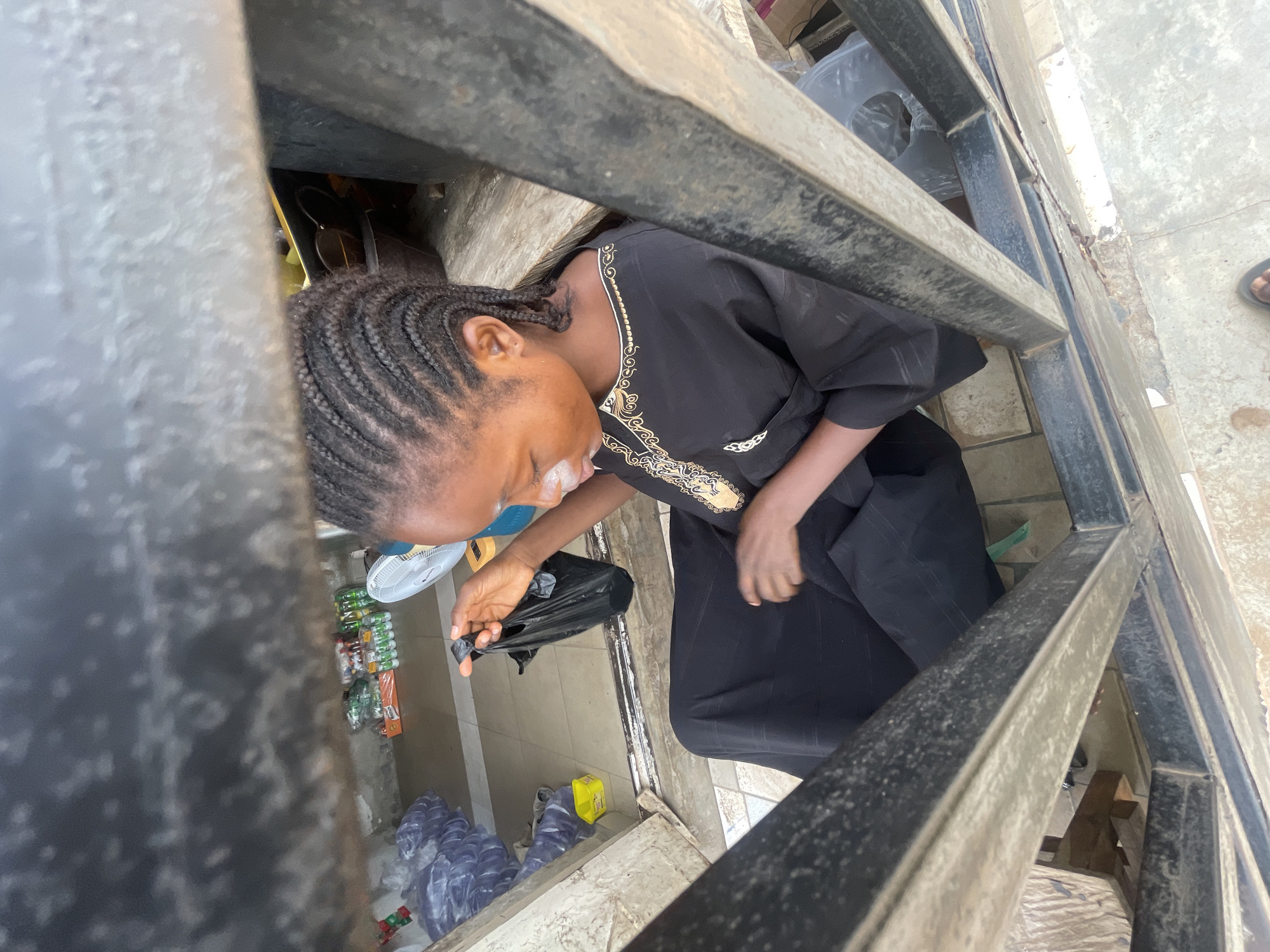 From a high, partially obstructed angle, a woman with braided hair is seen in Lokoja, Nigeria. She wears a dark, embroidered dress and appears to be reaching into a small shop space. The shop contains bottled drinks, sachets of water, and a fan. She holds a black plastic bag. The view is framed by a metal structure.
