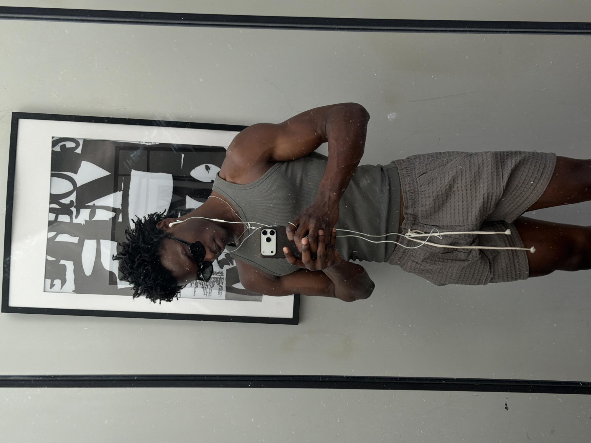 A reflection in a dirty mirror shows a man wearing a gray tank top, gray shorts, and sunglasses. He has dark curly hair and earbuds connected to a white smartphone he is holding. A framed black and white abstract artwork is on the wall behind him. The location is unknown and there is no indication that this photo was taken in London.
