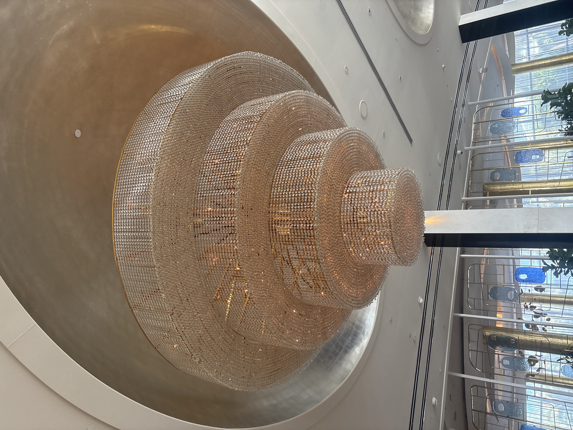 This image showcases a magnificent, multi-tiered crystal chandelier as the focal point of a grand, modern interior space, likely a luxury hotel or similar establishment in Miami Beach. The chandelier is massive, composed of countless small, clear crystal beads or prisms meticulously arranged in concentric, decreasing rings, forming a dazzling, dome-like structure. It emanates a warm, golden light from within, causing the crystals to sparkle brilliantly and cast intricate patterns.
The opulent light fixture is suspended from a large circular recess in a predominantly white or off-white ceiling. Parts of this recess exhibit a textured, reflective surface, possibly metallic, which catches ambient light. The ceiling also features subtle recessed lighting and sleek linear black accents.
To the right of the chandelier, the scene opens up to what appears to be a multi-story atrium or grand hall. Natural light streams in from large windows or a glass facade, through which hints of green foliage are visible, suggesting an outdoor view. The architectural elements in this section include clean white railings, horizontal gold-toned accents, and several irregularly shaped decorative panels in shades of blue and light blue glass or ceramic.
The overall aesthetic is one of modern elegance and luxury, characterized by clean lines, reflective surfaces, and a harmonious blend of natural and artificial light. There are no people or discernible activities, allowing the viewer to appreciate the architectural design and the impressive scale and craftsmanship of the chandelier. The bright natural light indicates it is daytime. No text is visible in the image.