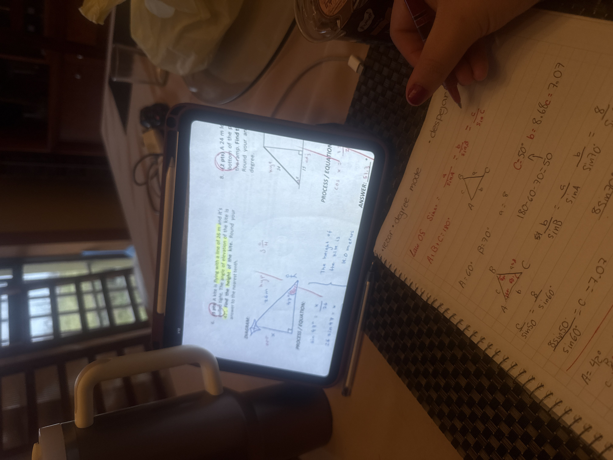 The image shows a person working on trigonometry problems. The person is holding a pen and writing on a notebook with trigonometry problems. The notebook includes diagrams of triangles and equations related to the Law of Sines. A tablet in front of the notebook displays trigonometry problems related to kites and ramps. A white mug is on the table in the bottom left corner.