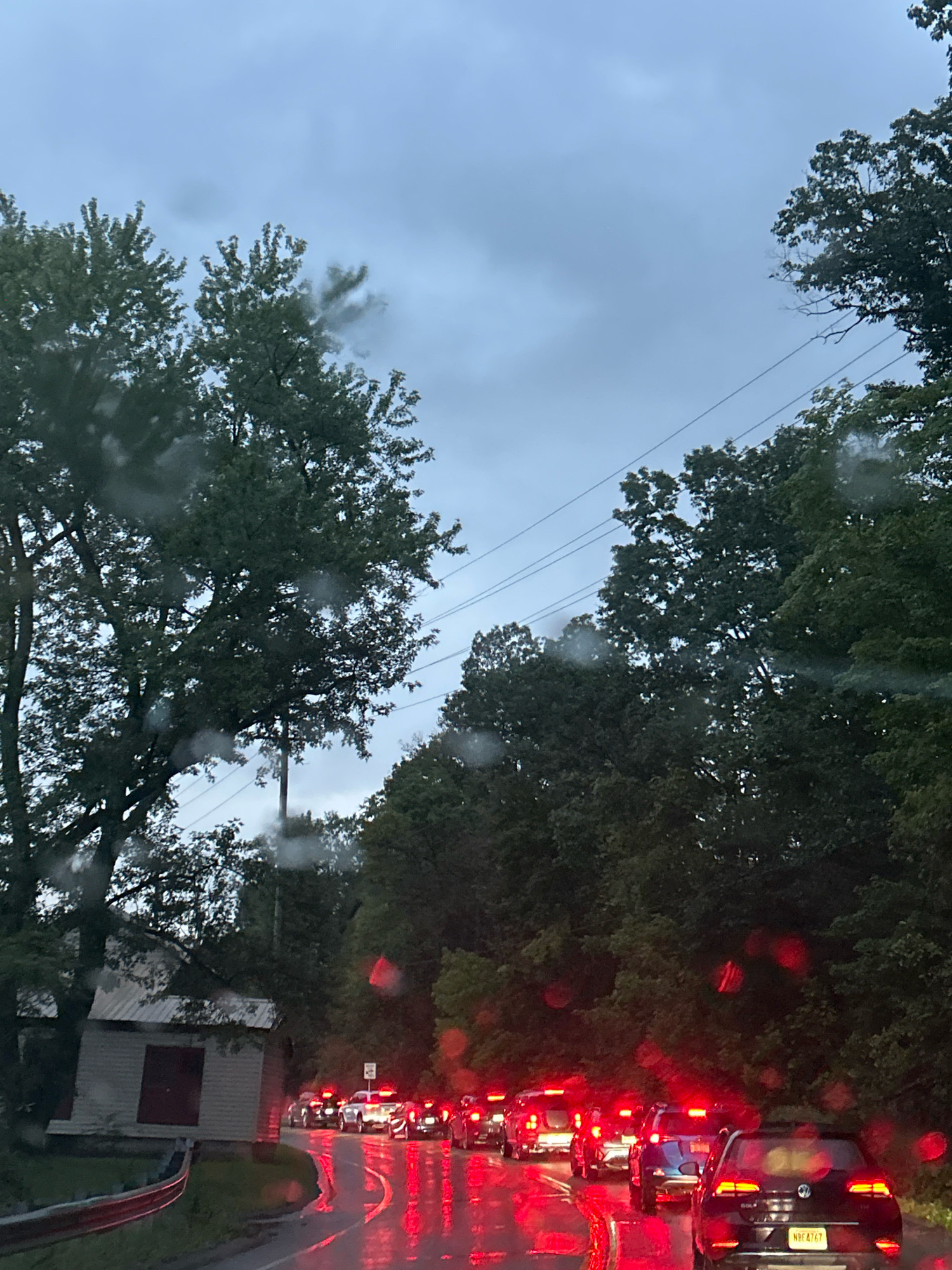 The image depicts a traffic jam on a wet, tree-lined road, likely during dusk or overcast weather. Raindrops are visible on the camera lens, creating red and white bokeh effects. A line of cars with their brake lights illuminated stretches into the distance. On the left side of the road, a small, weathered building with a red-doored garage is visible. The trees flanking the road are dense and green, and power lines can be seen strung between poles in the distance. The road surface is reflective due to the rain, mirroring the red brake lights of the vehicles. A license plate on the black Volkswagen in the foreground reads "NBE4767". Without additional context, it is impossible to determine the specific location within Pittsburgh, United States of America.