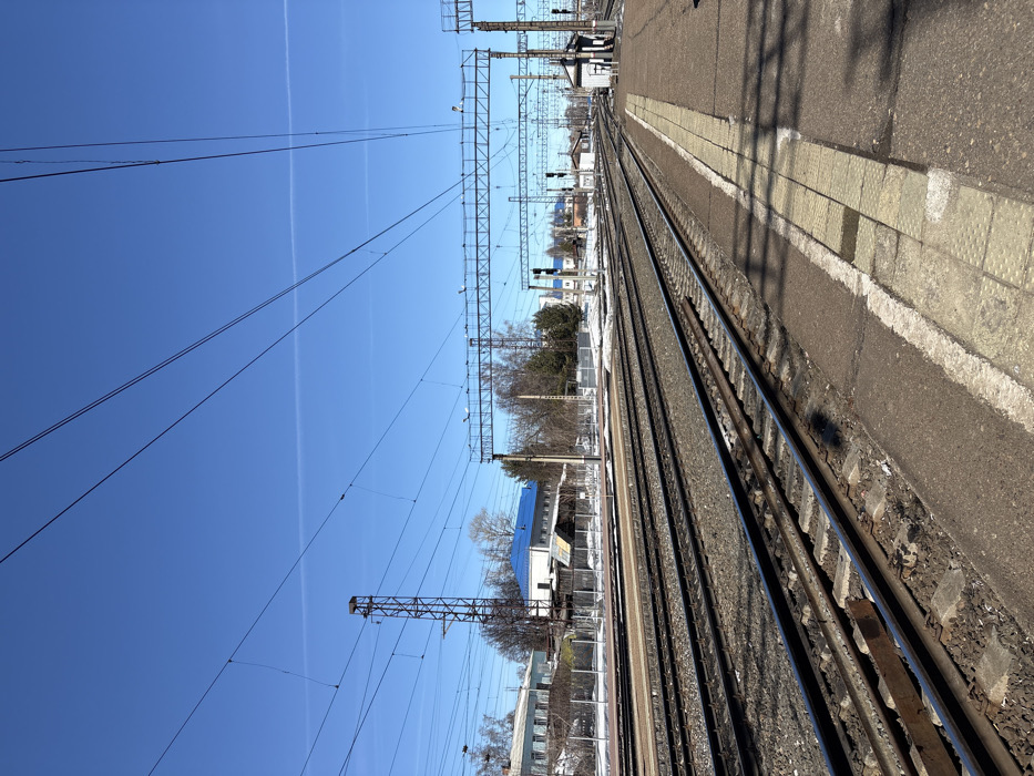 The image depicts a railway station in Kropachyovo, Russia, under a clear, bright blue sky with a single faint contrail visible. The scene is devoid of individuals. In the foreground, a concrete railway platform is visible on the right, featuring light-colored tactile paving blocks along its edge. Multiple sets of parallel railway tracks, approximately five visible, extend into the distance, with dark gravel ballast between them. Shadows from unseen structures and overhead lines are cast across the tracks and platform.
To the left of the tracks, in the midground and background, there are various buildings, including a structure with a distinctive bright blue roof and a multi-story building to its left. Bare trees and sparse brush are present behind a fence separating the tracks from these buildings. Patches of snow are visible on the ground beside the tracks and near the buildings, indicating a late winter or early spring setting. Overhead, a complex network of electrical catenary lines is suspended by metal gantries and poles. A tall, brown, lattice-style metal tower stands to the left of the tracks, possibly supporting signals or communication equipment. Further along the tracks on the right, a small white building, likely a signal box or utility structure, is present near the end of the platform. Several railway signals with dark casings and circular lenses are visible along the track lines in the distance.
