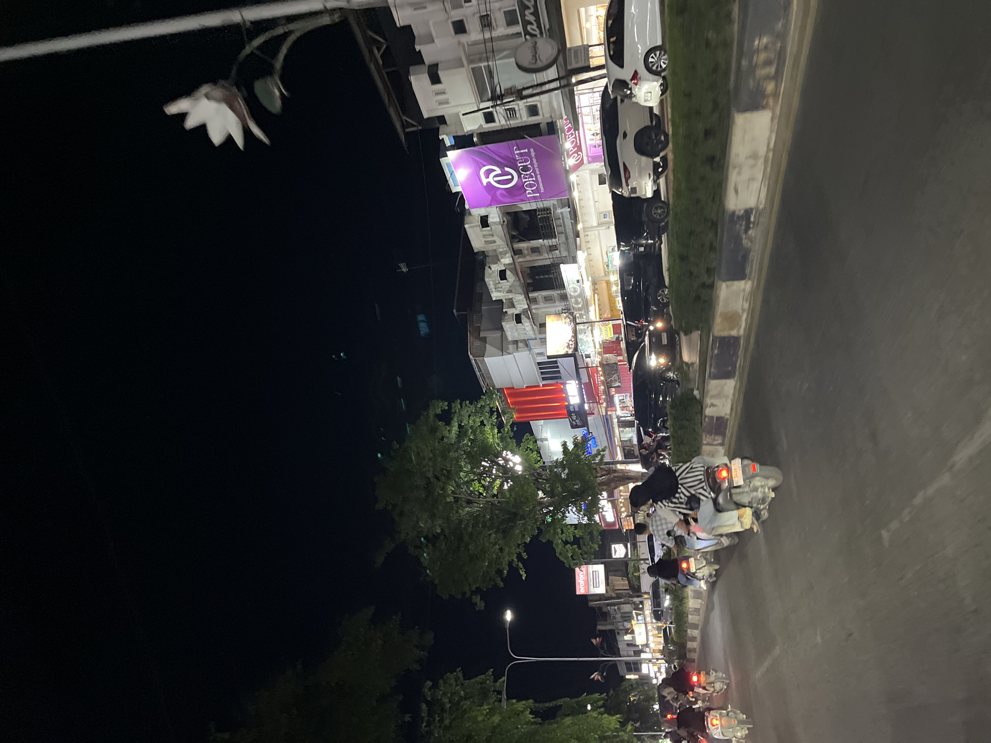 The image depicts a nighttime scene on a city street in Langsa, Indonesia. A road is visible in the foreground, with several motorbikes traveling in the same direction. On the right side of the road, there are parked cars, including a white SUV and a white sedan. Beyond the cars, there are multi-story buildings with various storefronts, illuminated by lights. One prominent sign displays "POECUT" in large purple lettering with a logo. Trees line the street, and a street lamp with a white floral ornament is visible in the upper right corner. The overall atmosphere suggests a bustling urban environment at night.