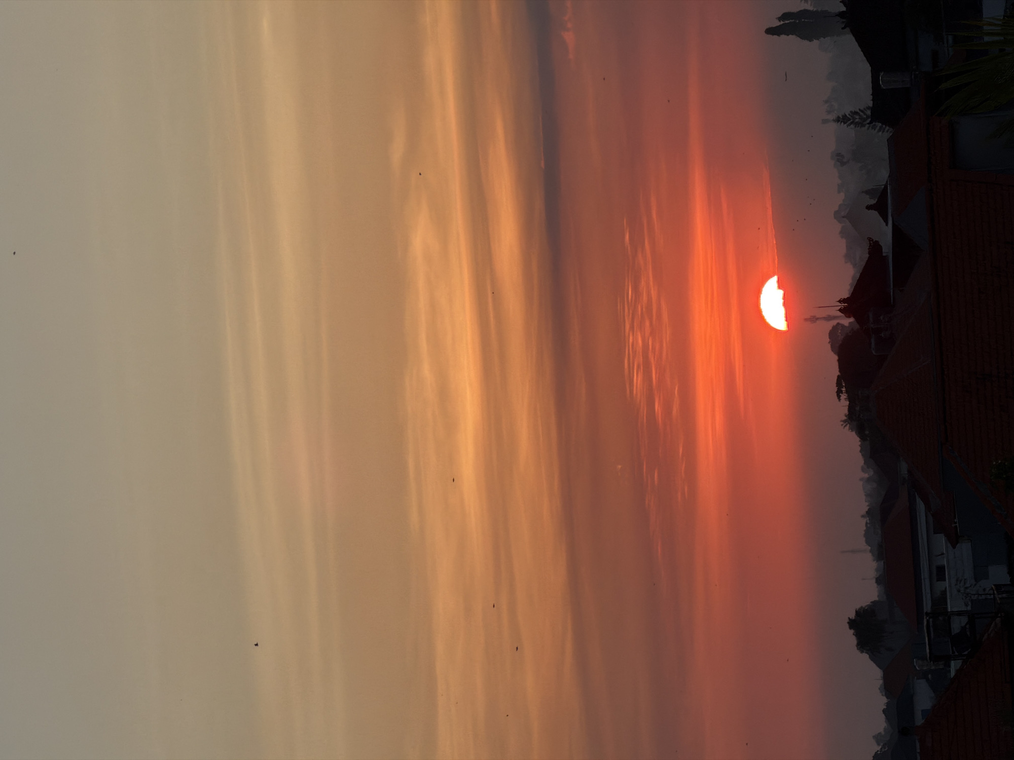 The image depicts a sunset over North Kuta, Indonesia. The sun is partially obscured by buildings and trees, casting a warm orange and red glow across the sky. The sky is streaked with clouds, ranging in color from pale yellow to deep orange and red. Silhouetted rooftops and trees are visible in the foreground, indicating a residential area. Small dark shapes, possibly birds, are scattered throughout the sky.