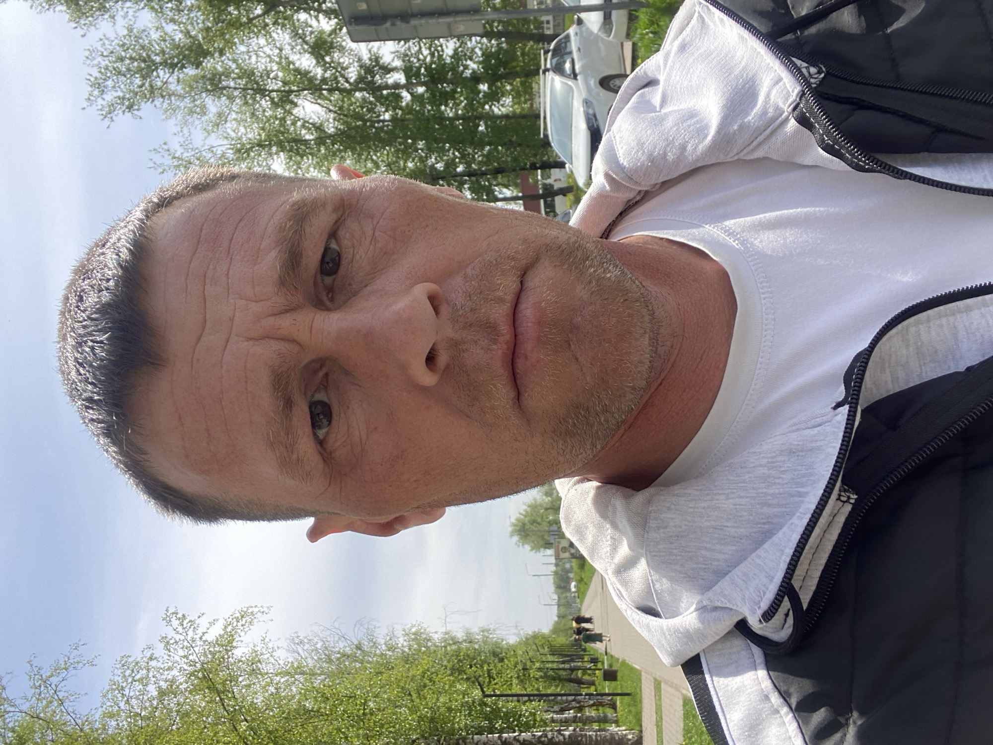 A close-up photograph shows a middle-aged man with fair skin and short, receding dark hair. He has light brown eyes and a salt-and-pepper beard. He is wearing a white t-shirt and a grey hooded sweatshirt under a black puffy vest.
The background includes green trees and a paved pathway suggesting a park or green space. There's a white car parked in the distance and what appears to be a utility pole.
The photo was taken in the city of Chaikovsky, Russia. The angle suggests the photograph was taken from a low vantage point, looking upwards.