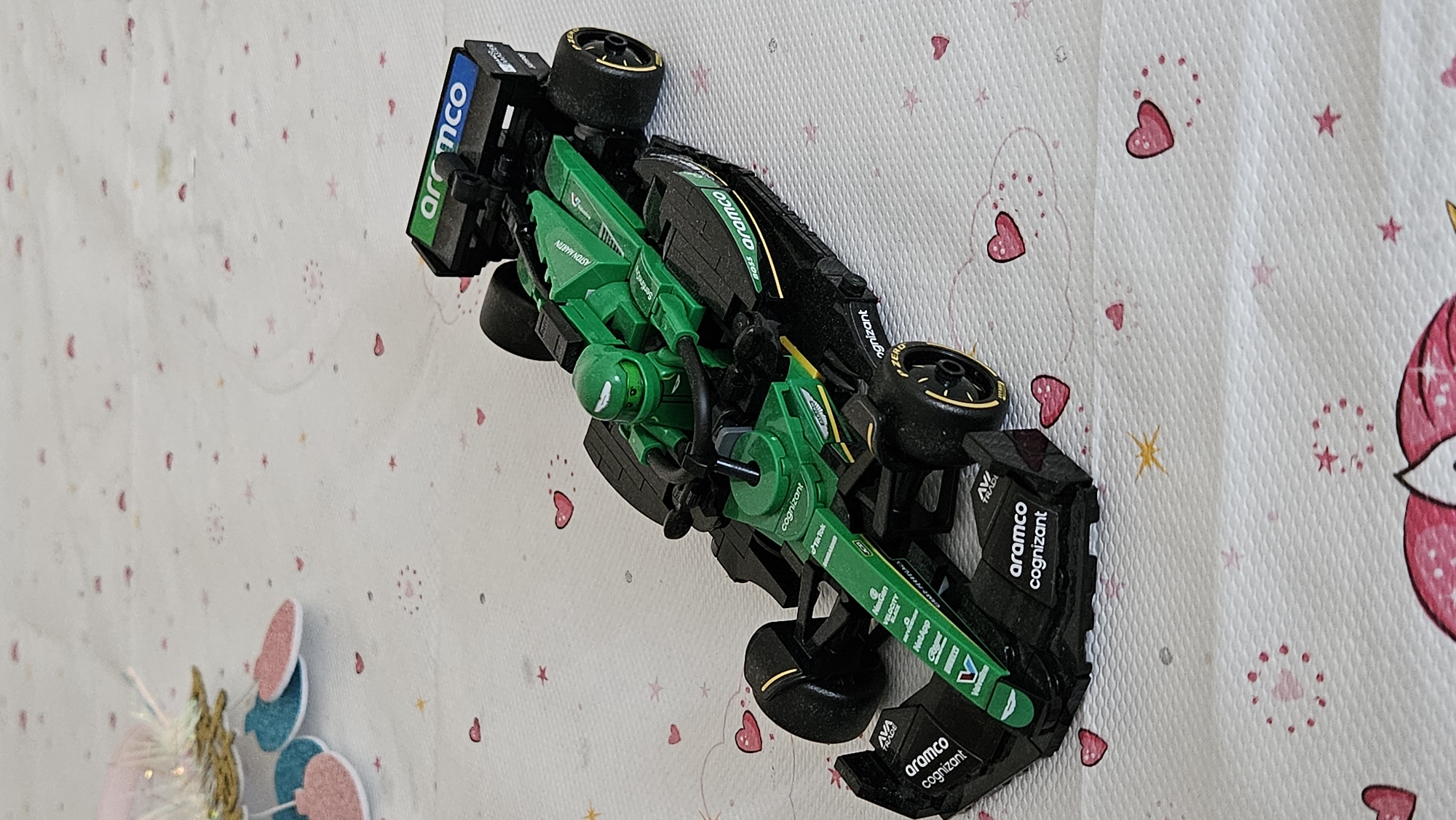 A green and black toy Formula 1 race car is displayed on a white tablecloth patterned with small pink hearts and stars. The car features sponsorship logos such as "aramco," "cognizant," and "ASTON MARTIN." The car is a detailed replica with a green driver figure in the cockpit. The tablecloth appears to be set on a flat surface, possibly a table, with a blurred background that includes decorative elements like glittery pink and teal ornaments and a white feathery trim. There is no indication of Bratislava, Slovakia, or any urban environment in the image.