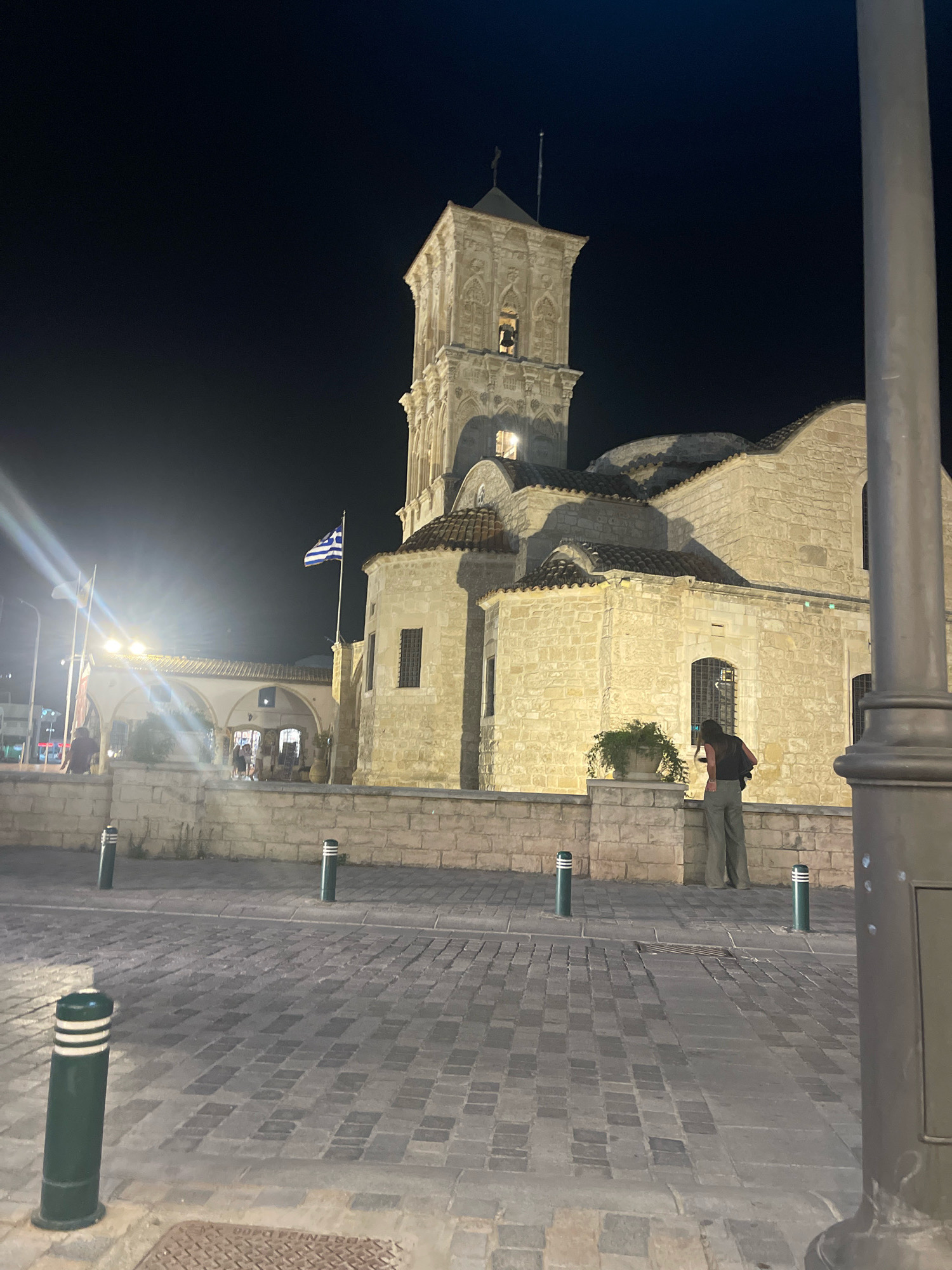 At night in Larnaca, Cyprus, the stone Church of Saint Lazarus stands illuminated. A tall bell tower with arched windows dominates the structure. In the foreground, a paved plaza is marked by dark green bollards with white stripes. A person wearing a dark shirt and green pants stands facing the church. To the left, a portico with arched openings provides access to shops. The Greek flag flies prominently on a flagpole. The sky is dark, indicating nighttime.