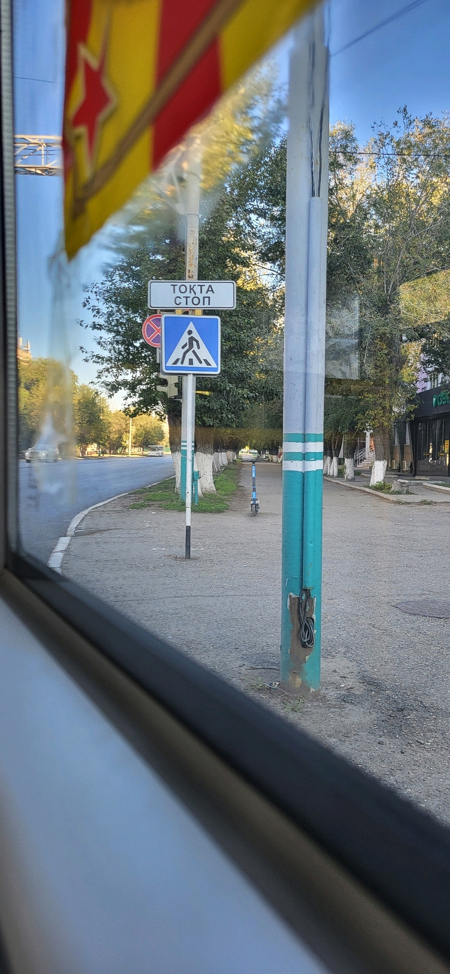 A reflection in a window shows a street scene with traffic signs and trees. In the foreground, a flag with red and yellow stripes and a red star is reflected. A sign with Russian text "ТОҚТА СТОП" (Tokta Stop) and a pedestrian crossing symbol is visible. To the left of this sign, a circular red and white prohibition sign is partially visible. A teal-colored pole with white stripes stands prominently in the mid-ground, with exposed wires at its base. The street is paved with asphalt, and a sidewalk runs alongside it, lined with trees whose trunks are painted white. A blue electric scooter is parked on the sidewalk further down the street. In the background, cars are driving on the road, and buildings are visible. The sky is clear and blue.