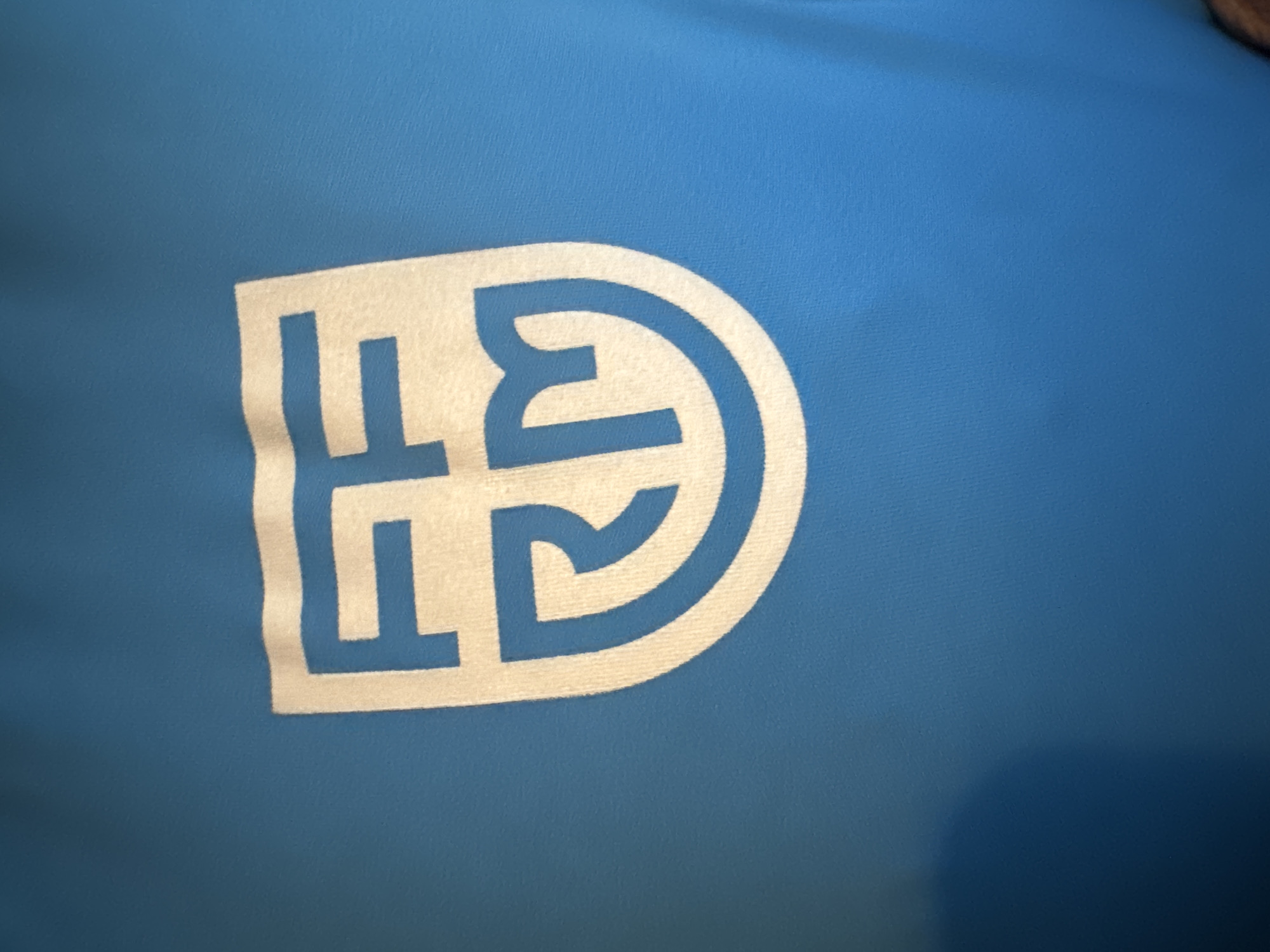 The image shows a white emblem on a blue fabric. The emblem is shield-shaped and contains stylized white letters. The top part of the emblem features two letters that appear to be "FF" or a similar angular design. Below this, a single letter "RM" is integrated into the lower portion of the shield. The fabric appears to be textured, possibly a t-shirt or athletic wear. There is a shadow casting on the lower left portion of the image, suggesting an external light source. There is no visible indication of the city of Murcia, Spain within the image itself.