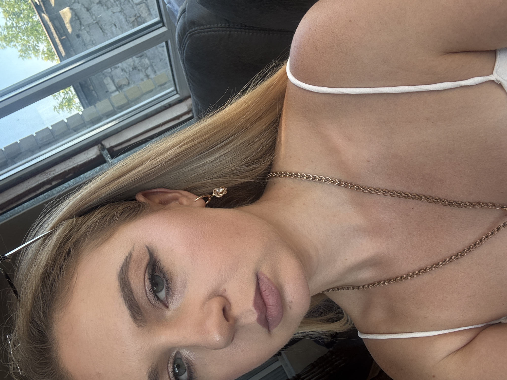 This is a close-up, sideways portrait of a young woman. She has blonde hair and is wearing makeup, including eyeliner and lipstick. She is also wearing a gold chain necklace and a gold earring. Her skin appears smooth and clear, and she is wearing a white top with thin straps. The background is blurred, but suggests an indoor setting with a window that looks out onto a brick wall and some greenery. The lighting suggests it is daytime. The woman's expression is neutral. There is no visible text in the image.