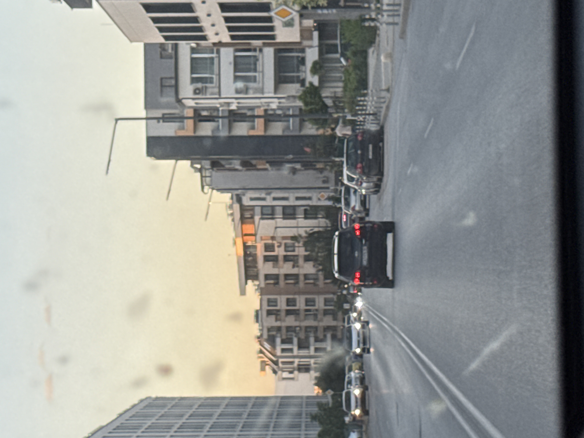 Captured through a window with noticeable spots, the image portrays a street scene in Sofia, Bulgaria. Tall buildings line the left side of the frame, varying in architectural styles. A row of cars moves along a multi-lane asphalt road on the right. The sky appears hazy and overcast, indicating potentially early morning or late afternoon light. Traffic appears moderate, with vehicles predominantly moving in one direction. No people are clearly identifiable in the foreground.