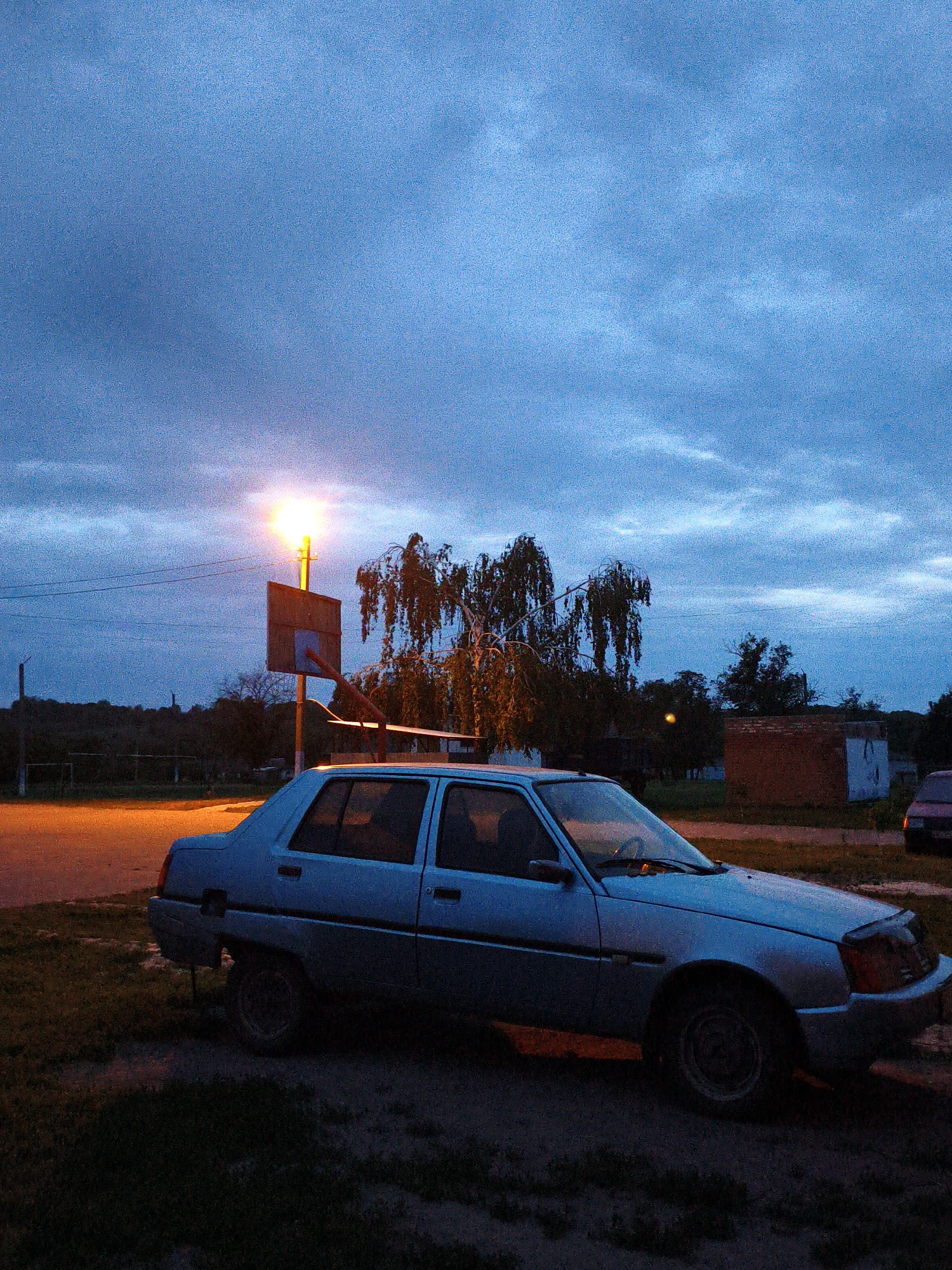 The image presents a quiet, dusk or early evening scene in Ustyvytsia, Ukraine. A light blue or silver sedan, possibly an older model, is the primary subject, parked on a dirt and grassy verge beside what appears to be a paved or dirt road. The car shows signs of age and use, with a slightly faded appearance.
In the mid-ground, a bright, warm-toned street light stands prominently, casting an orange glow. Directly beneath it is a basketball hoop with a plain, rectangular backboard. Behind the basketball setup, a large deciduous tree with weeping or trailing branches is illuminated by the street light, its form silhouetted against the darkening sky.
The sky is a deep, cloudy blue, indicative of twilight or early night, with visible cloud formations. In the background, a dense line of trees and foliage forms a dark horizon. To the right, a small, reddish-brown brick building or shed is discernible, and the partial outline of another parked car can be seen in the shadows.
The scene conveys a sense of stillness and calm, characteristic of a rural or small-town environment at the end of the day. There is no human activity depicted, emphasizing the quiet atmosphere. The artificial light from the street lamp creates a focal point against the encroaching darkness. No visible text is present in the image.