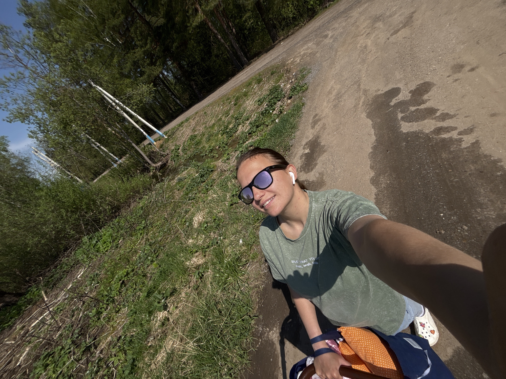 A woman takes a selfie outdoors, likely in Razdolye, Russia, based on the location request. She is wearing sunglasses, an earbud, a green t-shirt, shorts, and white Crocs. She is holding a bag that contains an orange polka-dot patterned item. The background consists of a dirt path, grass, and trees. Some utility poles are visible in the distance. The angle suggests she is holding the camera above her head. Sunlight is visible, casting shadows. There are patches of moisture on the ground.