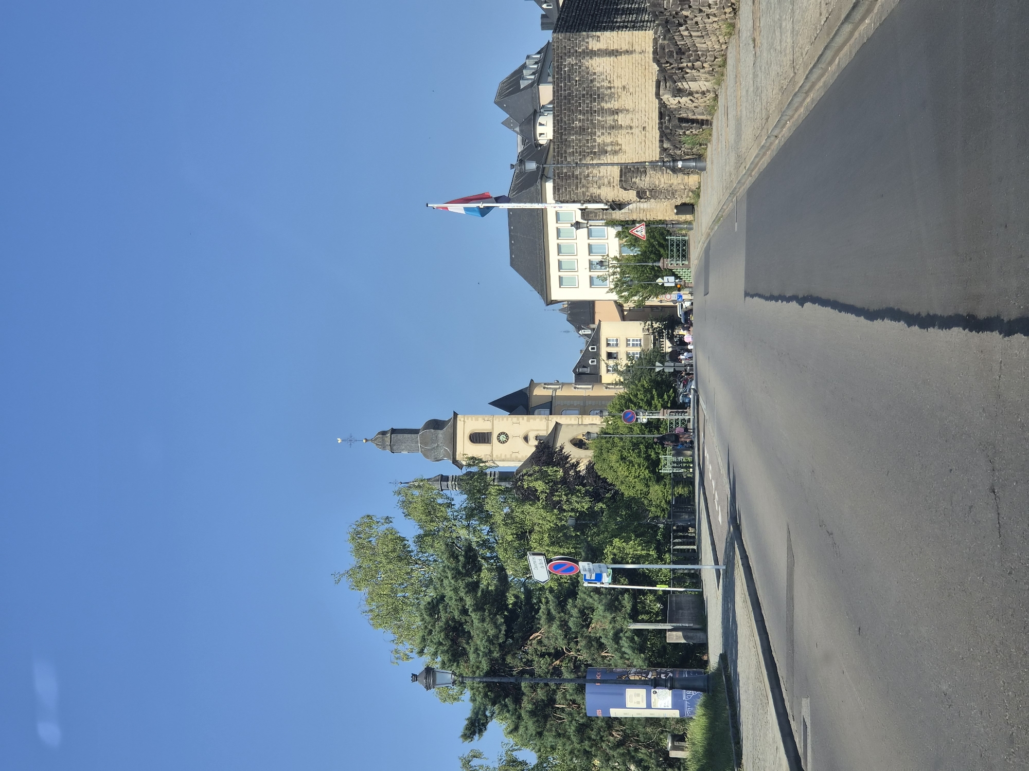 The image shows a street scene in Luxembourg City, Luxembourg. The street is paved, and there is a sidewalk on the right side of the road. On the left side of the street, there is a large tree and a lamppost. In the background, there is a church with a tall steeple, and other buildings, including a building with a large stone wall. A flag of Luxembourg is flying on a flagpole. There are a few cars and pedestrians visible on the street. The sky is clear and blue.