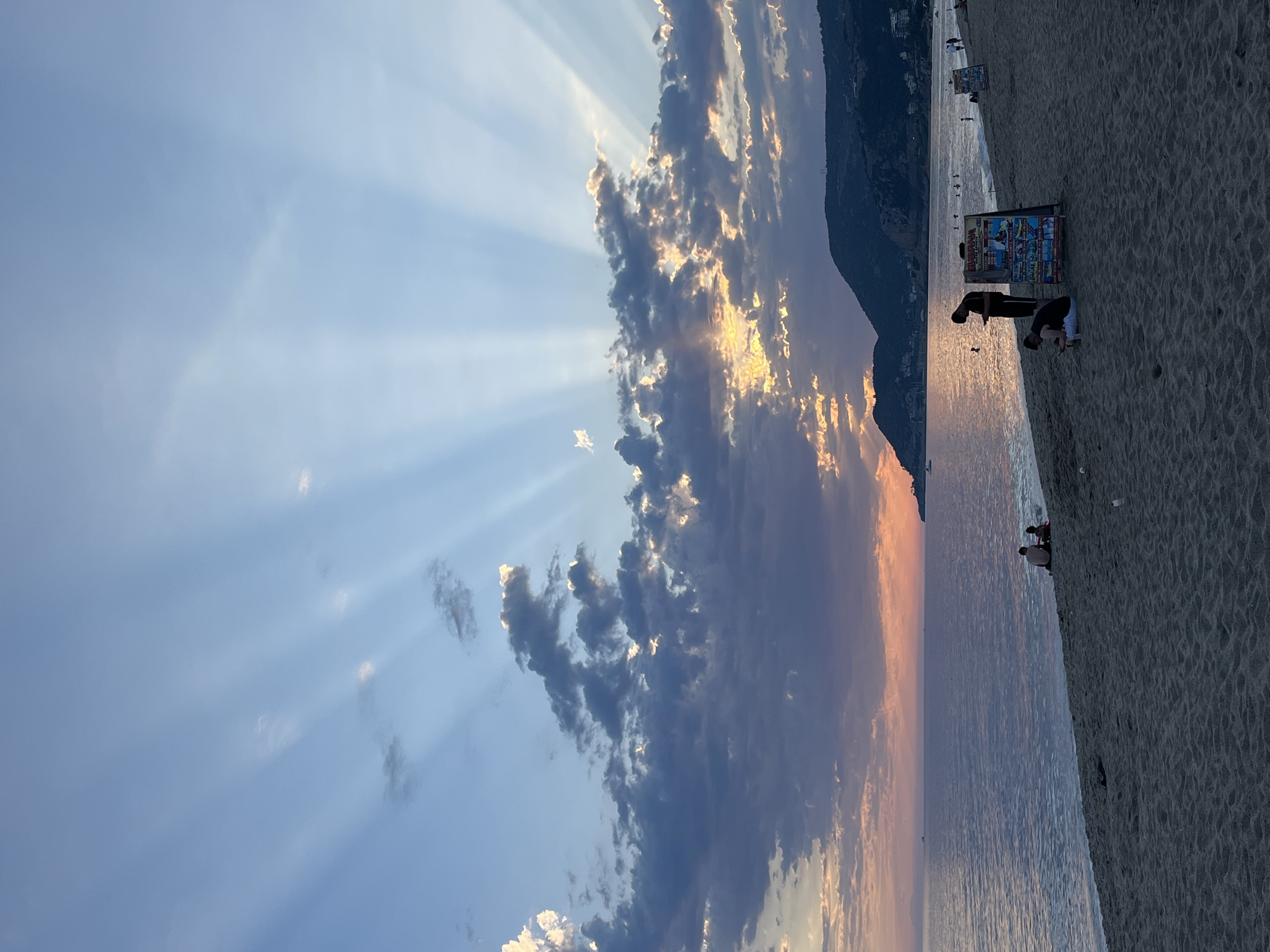 The image shows a beach in Alanya, Turkey, at sunset. The sky is a mix of blue and orange, with clouds and sun rays breaking through. The ocean is calm and reflects the colors of the sky. On the beach, there are people walking and sitting. There is a vendor with a display of goods. The mountains are visible in the background.