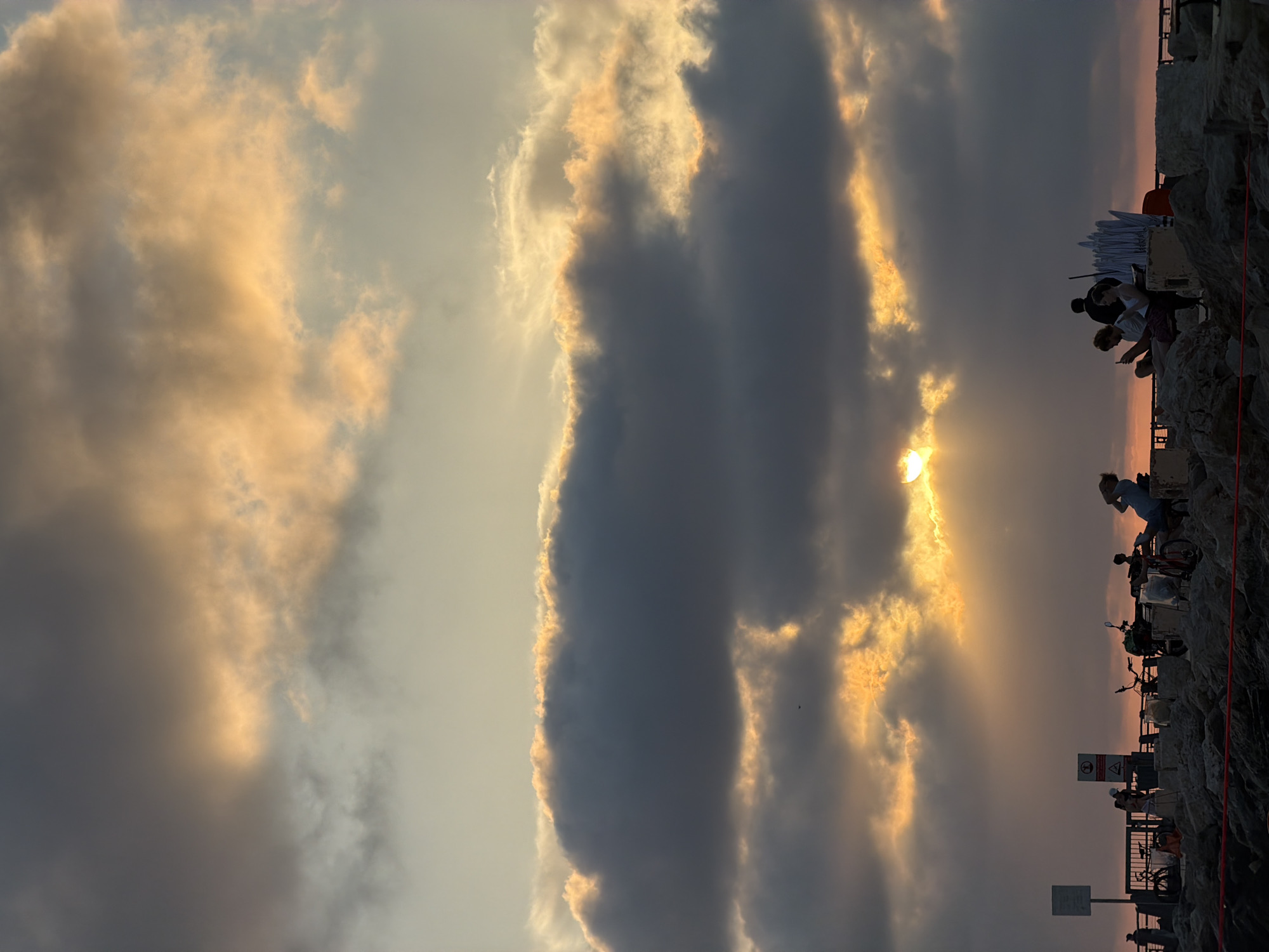 A long-shot, eye-level image shows a partially-cloudy sky near the ocean in Tel-Aviv, Israel. Silhouetted people stand near the coast. The sky is a mix of gray and orange, the sun partially visible through a break in the clouds. Along the coast, a rough stone wall runs parallel with a red rope fence. Sign posts and metal barriers are fixed on the wall, with people gathered at intervals, some sitting and others standing. Bicycles are parked nearby.