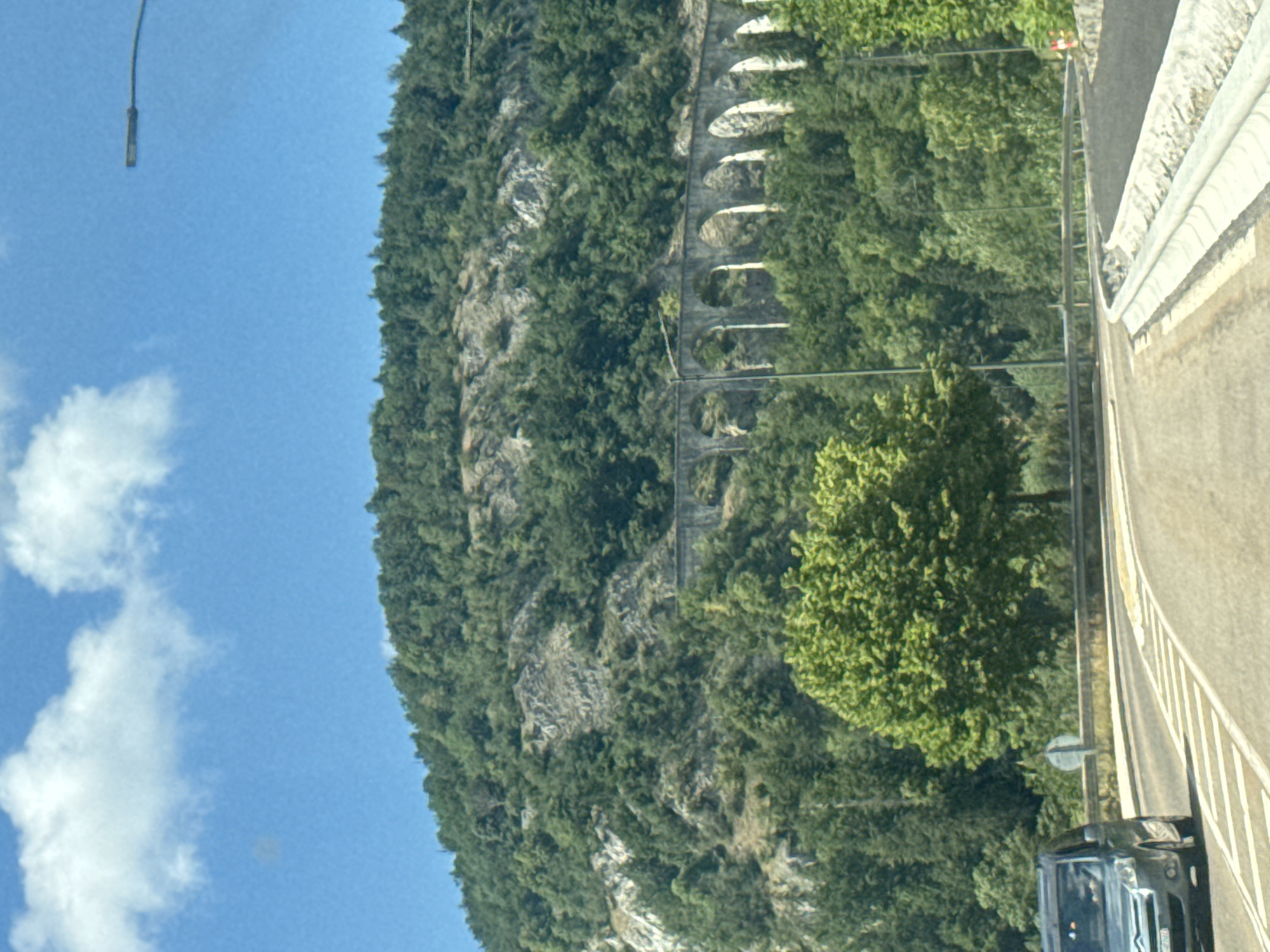 The image captures a scenic view of a mountainous landscape, likely in the region of Hauts de Bienne, France. In the foreground, a gray vehicle, possibly a van, is seen driving on a paved road. The road curves to the right, bordered by a concrete barrier on the right side. In the middle ground, a distinctive stone aqueduct with multiple arches traverses the hillside. The aqueduct is surrounded by lush green trees. The background is dominated by a steep, tree-covered mountain, with rocky outcrops visible. The sky above is blue with scattered white clouds, and a street lamp is visible on the right side of the frame. The overall impression is one of a natural, somewhat rugged environment integrated with historical infrastructure.