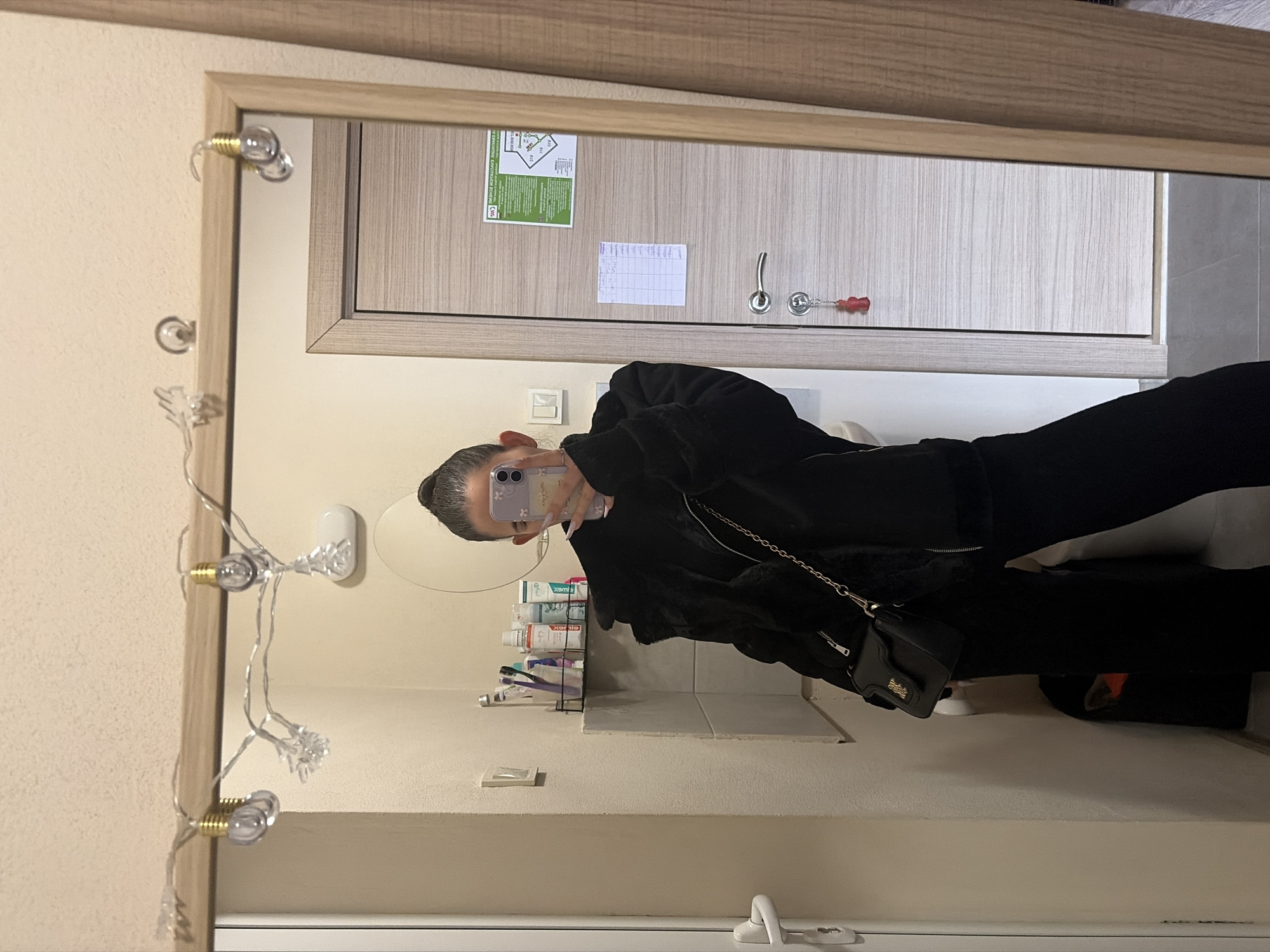 The image shows a mirror reflection of a person taking a selfie in a bathroom. The person is wearing black clothing and holding a phone with a lavender phone case. The bathroom has a sink, a rack with toiletries, and a string of light bulbs framing the mirror. In the reflection, a closed wooden door with a lock and a map-like poster is visible. Based on the style and features of the interior, the location is likely a hotel room or apartment in Sofia, Bulgaria.
