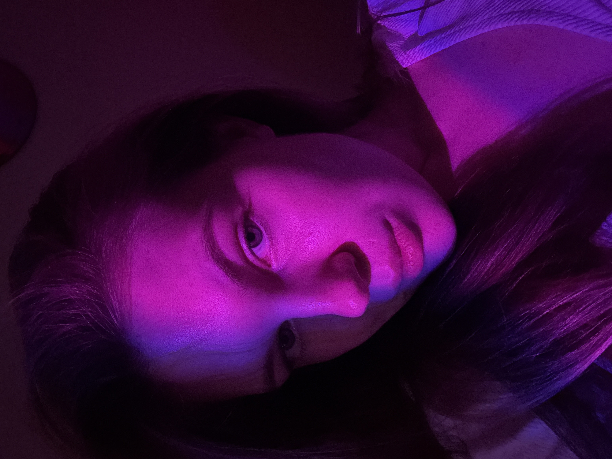 A close-up, rotated portrait of a fair-skinned person is illuminated with purple and magenta lighting. The person's face, including one visible eye, nose, and lips, is in focus. Dark hair surrounds the face. The person is looking slightly downward. The background is obscured by shadows and the strong colored lighting. No location information can be determined from the image.