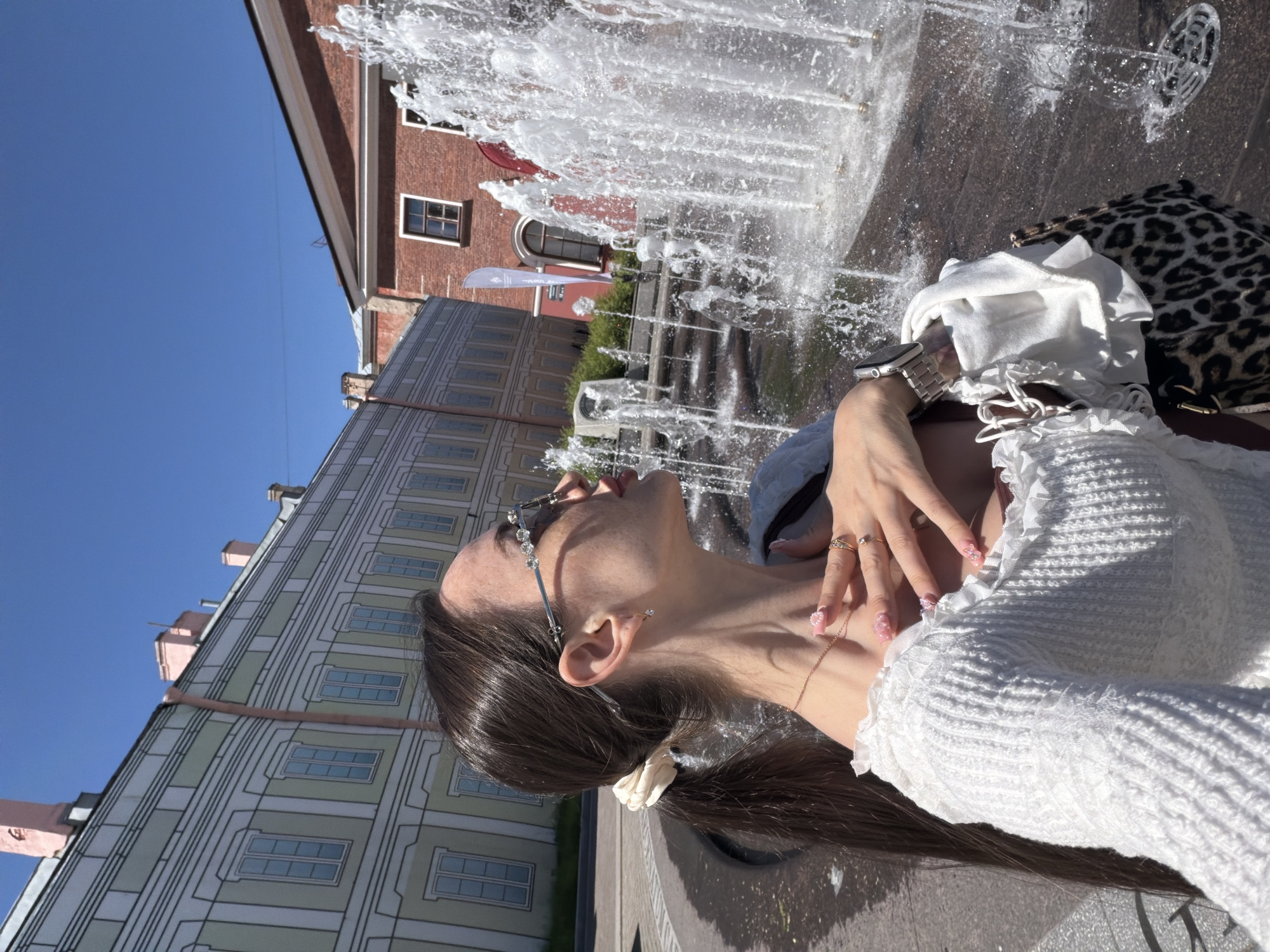 The image depicts a female individual in Saint Petersburg, Russia, standing outdoors on a clear, sunny day near an active water fountain. The woman is positioned in the foreground, facing right with her head tilted upwards, her gaze directed towards the upper right. She has long, dark brown hair secured in a low ponytail with a light cream-colored scrunchie. She wears sunglasses with decorative clear gems along the top edge of the frames. Her attire includes a white, long-sleeved, ribbed knit top with ruffled details at the neckline. A thin gold chain necklace with a small pendant is visible around her neck. Her right hand rests gently on her upper chest, revealing pink nail polish with small decorative elements and multiple gold-toned rings on her fingers. A silver-toned watch with a metal link band is visible on her left wrist. She carries a leopard-print shoulder bag, from which a folded white garment is partially visible.
To the right of the woman, a multi-jet fountain is actively spraying water upwards, creating white plumes and visible droplets against a dark paved surface. In the background, under a cloudless blue sky, two distinct buildings are visible. On the left is a long, light olive-green or pale yellow building with white architectural trim and trompe l'oeil painted windows, featuring light pink chimney-like structures on its roofline. To the right, a reddish-brown brick building with dark windows and white trim is partially visible, with a tall white flag or banner affixed to its facade near the fountain.