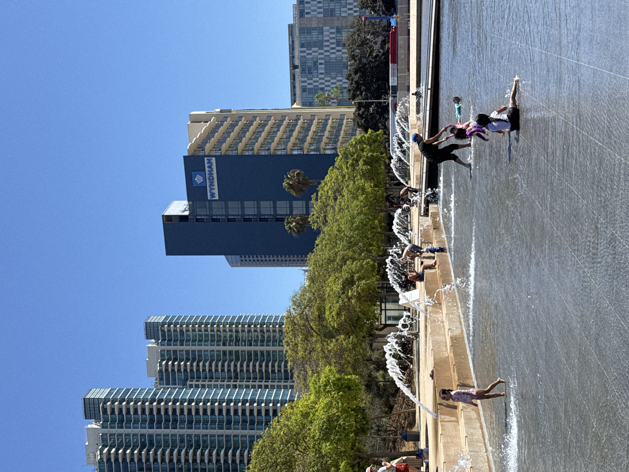 The image depicts a sunny day at a waterfront urban park in San Diego, California, USA. In the foreground, a group of children are playing in a shallow water fountain area. One young girl in a floral swimsuit stands in the water, while another child sits on the wet pavement. A woman, likely an adult, is interacting with two children in the fountain; she is bending down, possibly assisting them or playing. Further back, other individuals are visible, some sitting and observing, and at least one child is lying on their stomach in the water.
The midground of the scene features lush green trees and a raised seating area with tiered concrete steps. Fountains are actively spraying water, creating a dynamic and refreshing atmosphere. Behind the park, the cityscape rises with several modern high-rise buildings. Prominently, a blue building with "WYNDHAM" branding is visible, along with other tall structures with balconies and many windows. The sky is a clear, bright blue, indicating good weather. The overall environment suggests a public space designed for recreation and leisure, popular on a warm day.