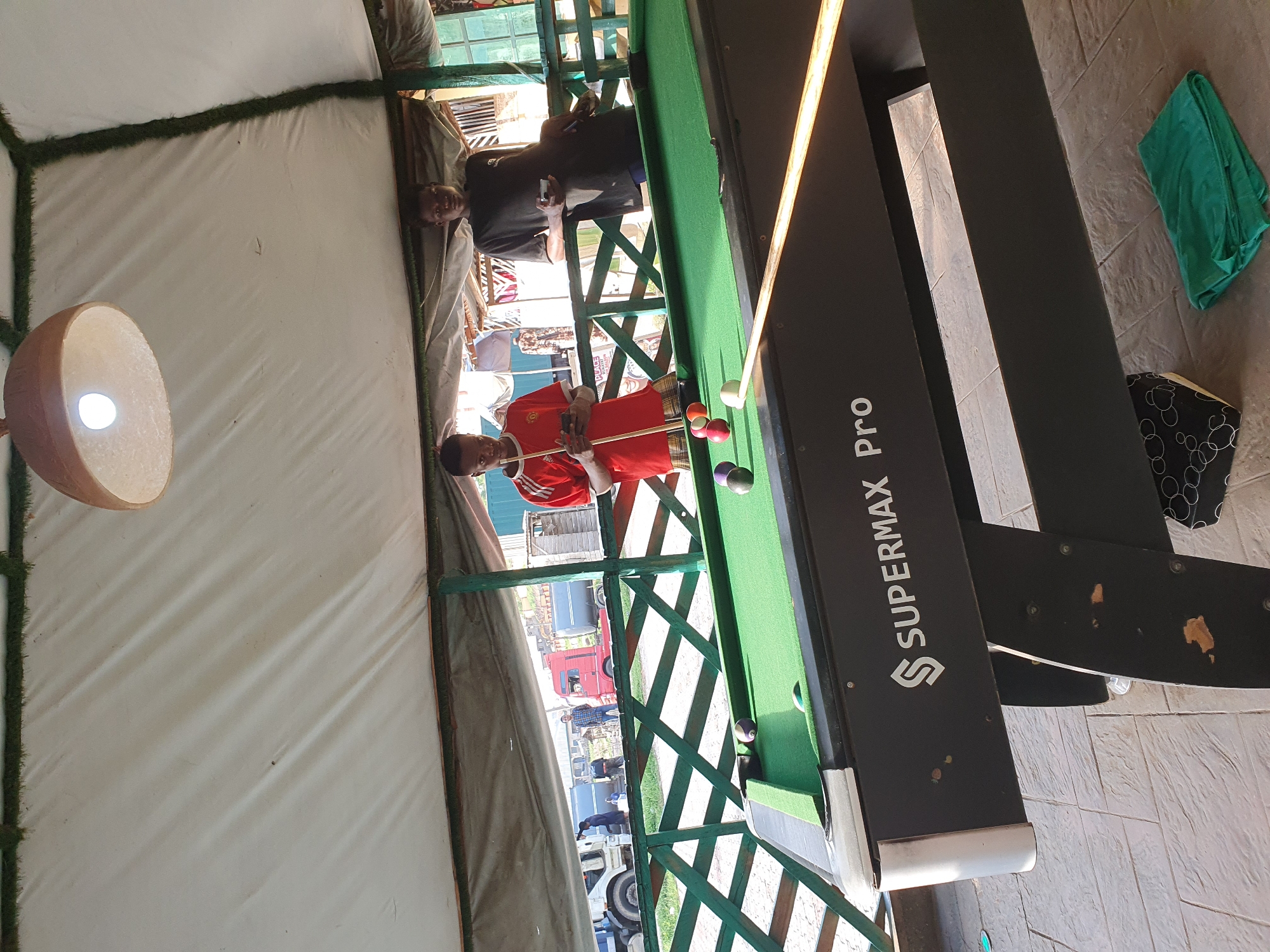 The image depicts an outdoor billiards area under a white tent. Two young men are present. One, wearing a red t-shirt with white stripes and plaid shorts, stands near the billiards table, holding a cue stick and looking at his phone. The other, in a black t-shirt, leans against a railing and also holds a phone. The billiards table, labeled "SUPERMAX Pro," has several balls on it, including a cue ball and colored balls. In the background, several trucks and a wooden structure are visible, suggesting a commercial or roadside location within Lokoja, Nigeria. The ground is paved with textured tiles. A green cloth is folded on the ground.