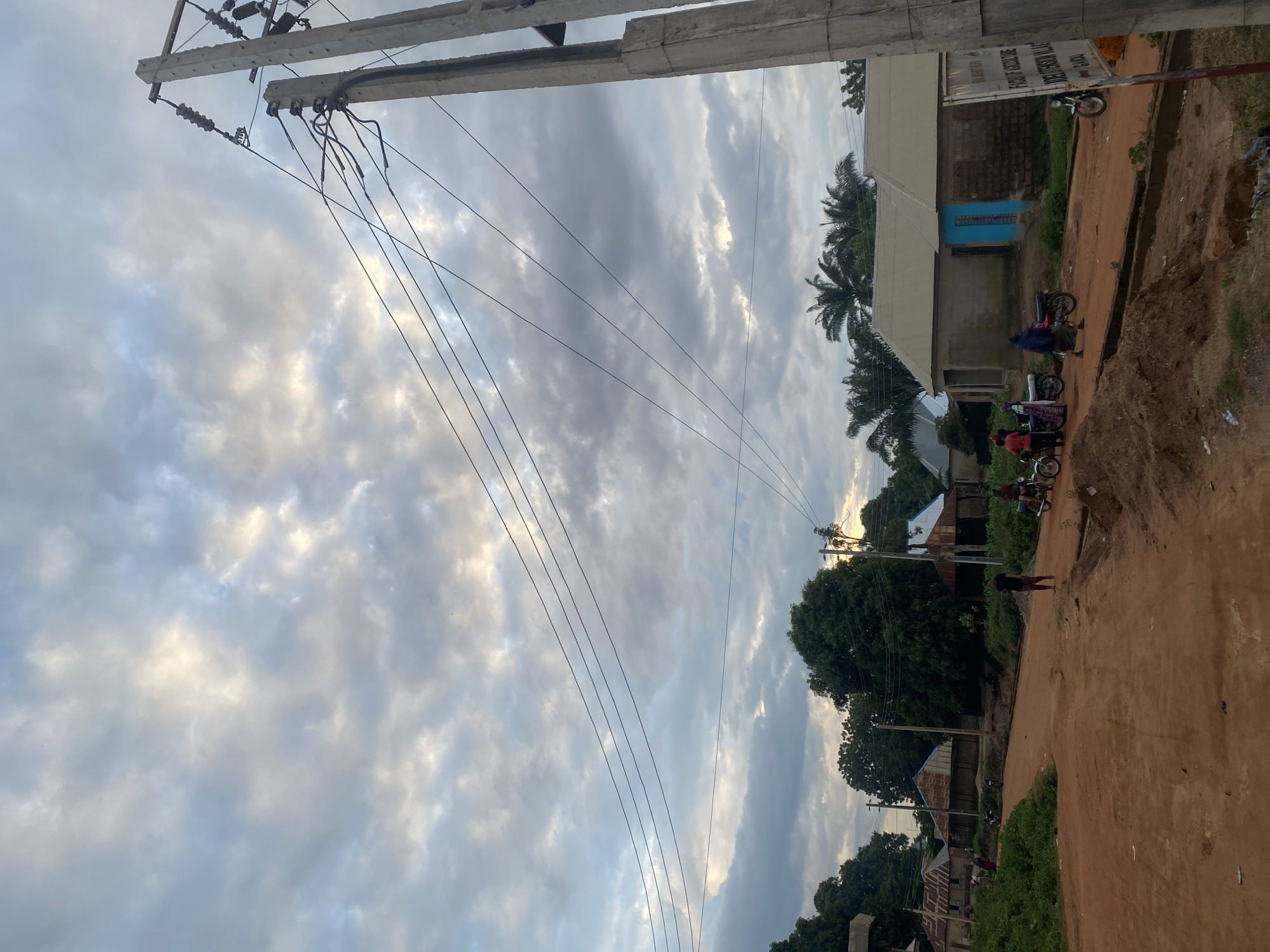 The image depicts a dusty, unpaved road in a rural area, possibly a village in Lafia, Nigeria. The sky is overcast with dramatic clouds, suggesting it might be late afternoon or early morning. Several modest buildings with corrugated metal roofs are visible on either side of the road, some with brick walls and others with what appears to be mud or plaster. A prominent concrete utility pole with numerous wires stands on the right side of the frame, dominating the foreground.
There are a few people present. On the left side, near some buildings, a small group of individuals and several motorcycles are gathered. In the middle of the road, a man in a black shirt and maroon pants stands. Further down the road on the right, another man is seen riding a motorcycle. The ground is a reddish-brown dirt, with some piles of excavated soil or debris along the roadside. Green vegetation, including trees and bushes, is scattered throughout the scene. A signpost is visible on the right, partially obscured, with text that appears to be in English and possibly another local language. The overall impression is one of a quiet, working village community.