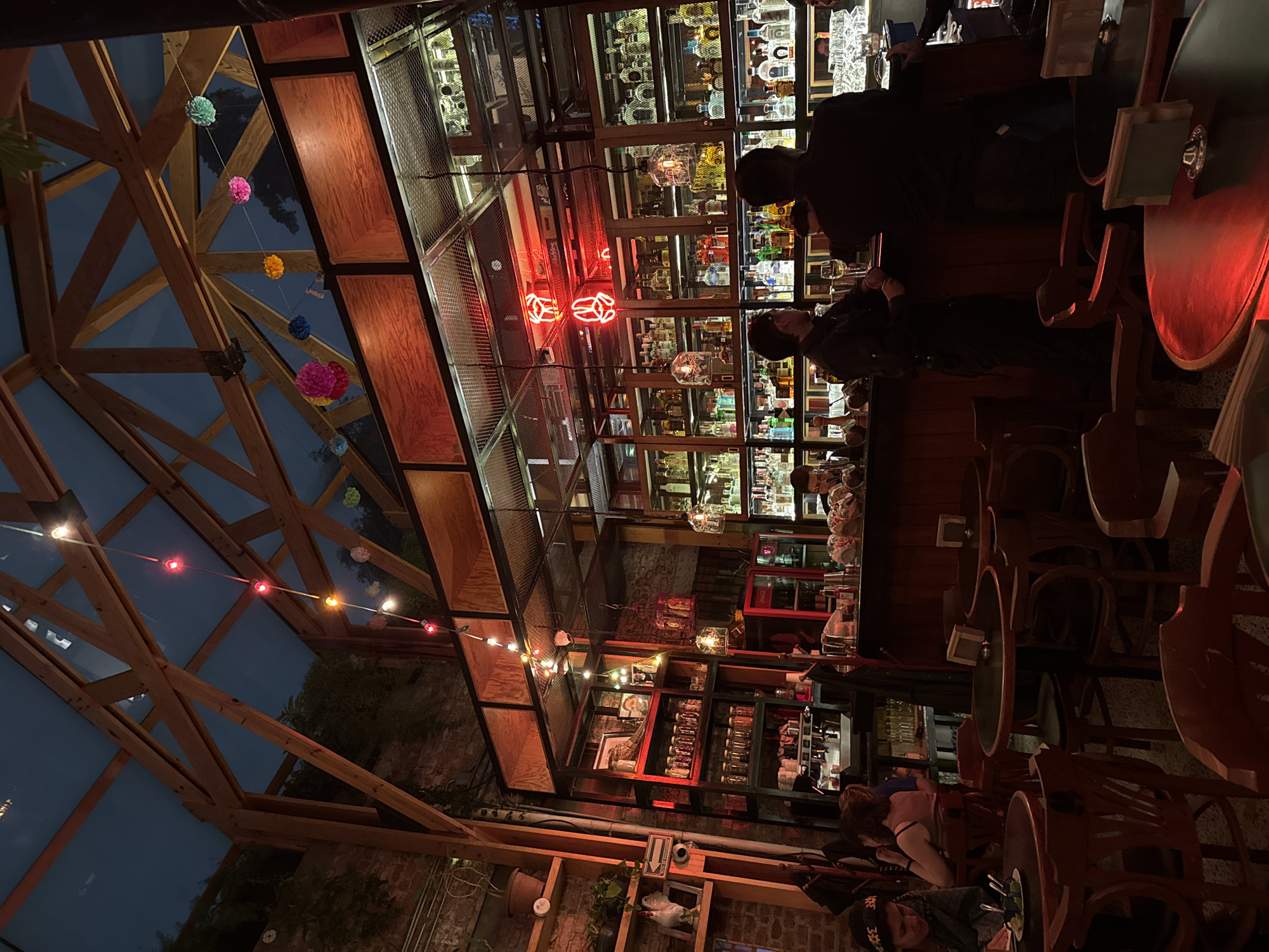 The image captures the interior of a bar or restaurant, likely in Mexico City, Mexico, characterized by its atmospheric lighting and unique design. The location seems to have a garden or outdoor seating area that is covered and surrounded by what seems like a wooden frame.
In the foreground, tables and chairs are visible, partially obscured by the low lighting. Patrons are seated, one person with a distinctive bandana. The bar itself is centrally located in the image, stocked with numerous bottles of liquor and glasses. A neon sign, shaped like a pair of wrenches, adorns the bar area, contributing to the vibrant ambiance. Overhead, strings of colorful lights and paper decorations are strung across the wooden beams of the structure. The building's design incorporates exposed brick and wooden structures, lending a rustic charm.
The overall scene is bustling with activity, illuminated by a mix of ambient lighting and neon signs. The shelves behind the bar are stocked with a large variety of beverages. The image is captured from a slightly tilted perspective, providing a dynamic view of the space.