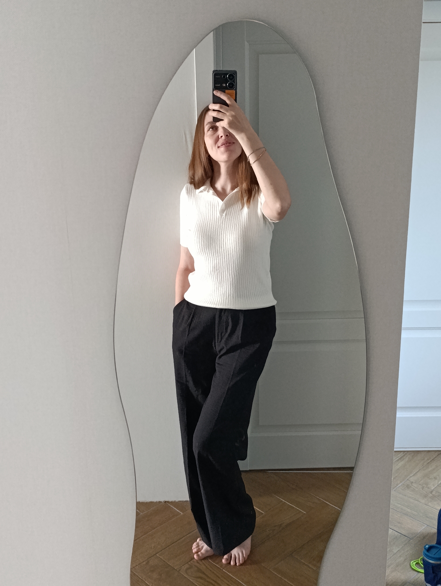 A woman is taking a selfie in a mirror in what appears to be an indoor setting, potentially a residential apartment. The woman is fair-skinned with reddish-brown hair and is wearing a short-sleeved, ribbed white top and loose-fitting black pants. She has one hand raised, obscuring part of her face while holding a smartphone. She appears to be barefoot.
The mirror is irregular in shape with a light-colored frame and reflects a portion of a room with light-colored walls, white door frames, and a wood-paneled floor.
The location, Chaikovsky, Russia, is mentioned. The presence of the mirror in a residential apartment and other details is inferred based on context and observations in the image.