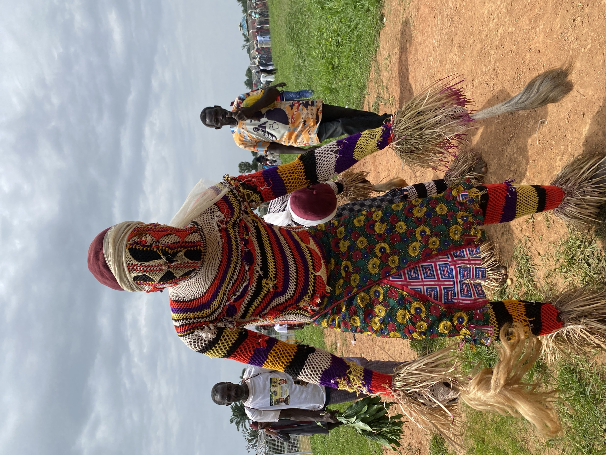 The image shows a person in a colorful, elaborate costume with straw-like elements on the hands and feet, standing in an outdoor area in Takau, Nigeria. The costume consists of patterned and striped sections in red, yellow, purple, and black. The person's face is covered with a patterned mesh. The location appears to be a field with grass and red soil. There are people in the background, including one in a patterned shirt and another holding leaves. The sky is overcast.