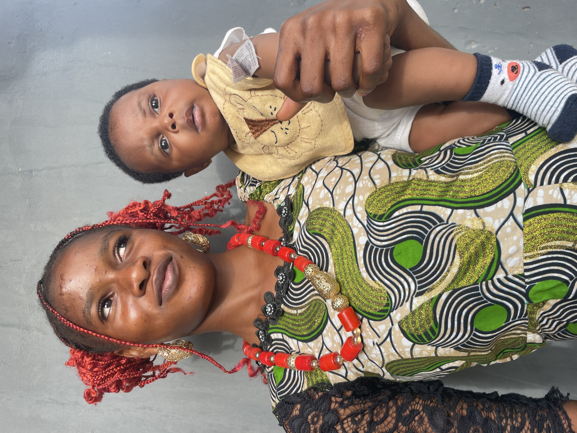 This vertical portrait features an adult female and a baby, likely a mother and child, posed as if lying down with the baby resting on the adult's chest or stomach. The adult female is positioned at the bottom, looking upwards and slightly to her right, while the baby is on top, gazing directly at the camera.
The adult female has dark skin and red braided hair or extensions styled around her face. She wears ornate gold earrings and a prominent red and gold beaded necklace. Her attire is a vibrant traditional African print dress, often called an ankara, with a rich pattern of green, black, and white swirls, interspersed with glittering gold details. Black lace forms part of the dress's design. Her expression is soft and contemplative.
The baby has dark skin and short, dark hair. They are dressed in a light-colored onesie and a yellow bib featuring an embroidered cartoon lion's face. Their feet are covered with white and blue striped socks adorned with an animal face pattern. An adult hand is visible, gently holding the baby's arm. The baby's expression is neutral and attentive.
The background is a plain, light-grey surface, suggesting an indoor setting. The traditional African attire and accessories worn by the adult female align with the provided location context of Badagry, Nigeria. There is small, unreadable text on a tag attached to the baby's clothing.