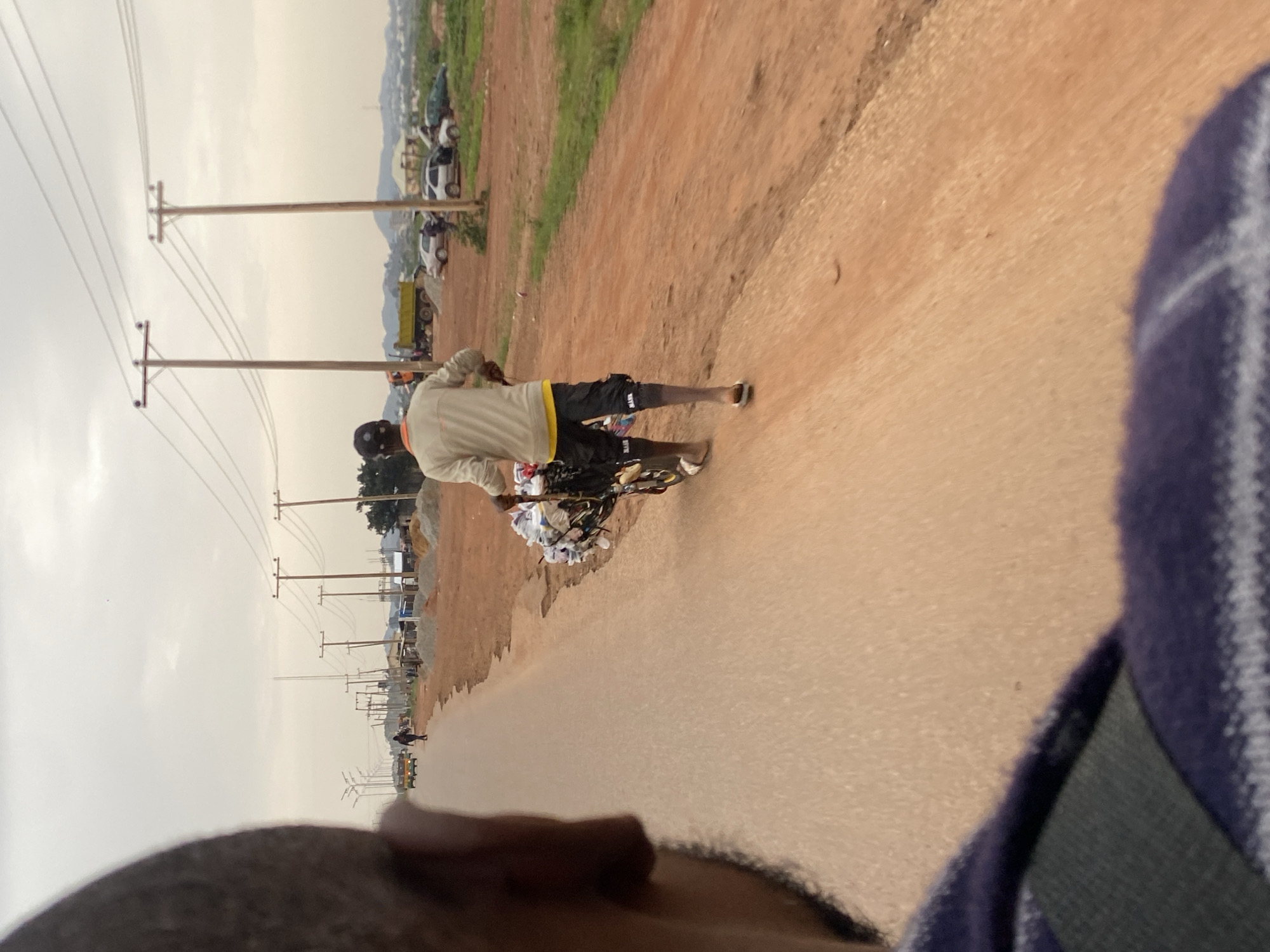 An outdoor, medium-shot, eye-level, wide-angle photograph captures a man walking on the right side of a paved road in Jos, Nigeria. The man is seen from behind, wearing a light-colored long-sleeved shirt over a yellow t-shirt, black shorts, and flip-flops. He is pushing a cart laden with various items, including what appear to be plastic bottles and bags. His left leg is bare from the knee down.
The road is primarily tan-colored asphalt, with a dirt shoulder on the right. In the distance, several utility poles with overhead wires stretch across the frame. Beyond the poles, the landscape consists of a mix of dirt construction areas with piles of sand and gravel, a few parked vehicles, and some buildings. Hills can be seen in the far background under an overcast sky.
In the foreground, a person's head and shoulder are visible in the lower-left corner, suggesting the photograph was taken from a moving vehicle. The person's head is partially blurred, and they are wearing a purple and white striped garment. The overall scene depicts a typical day in a developing urban environment, possibly a construction or industrial zone on the outskirts of Jos.