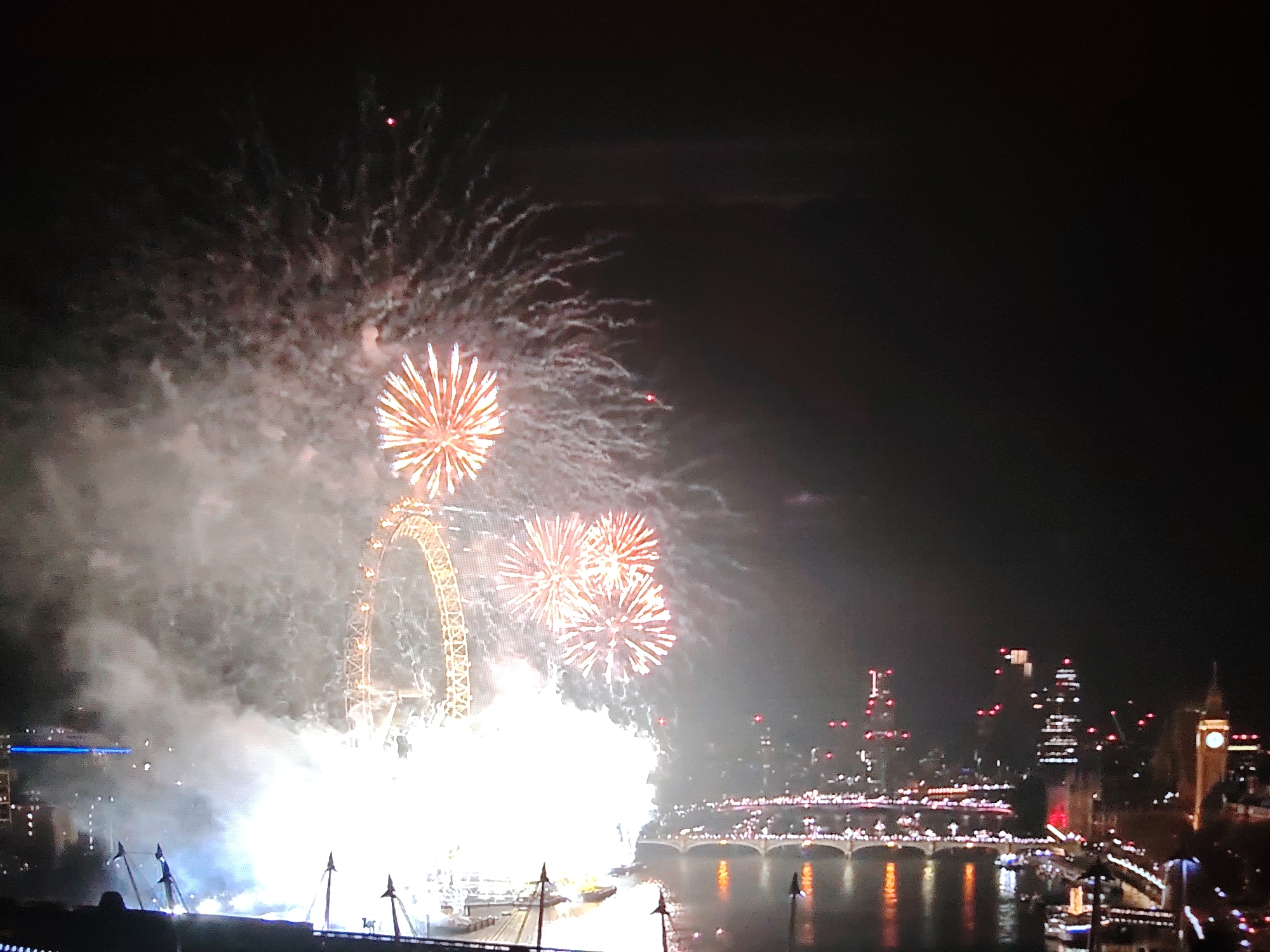 The image depicts a nighttime fireworks display over a city skyline, with the London Eye prominent in the foreground. The London Eye, a large Ferris wheel, is illuminated and surrounded by bright explosions of fireworks, primarily in shades of orange and white. Smoke from the fireworks drifts across the scene. In the background, a cityscape with illuminated buildings and a bridge spanning a body of water is visible. A clock tower, resembling Big Ben, can be seen on the right side of the skyline. The water reflects the lights from the city and the fireworks. The overall scene suggests a celebratory event. The location is the city of London, United Kingdom, specifically along the River Thames. There are no discernible people or specific interactions visible.