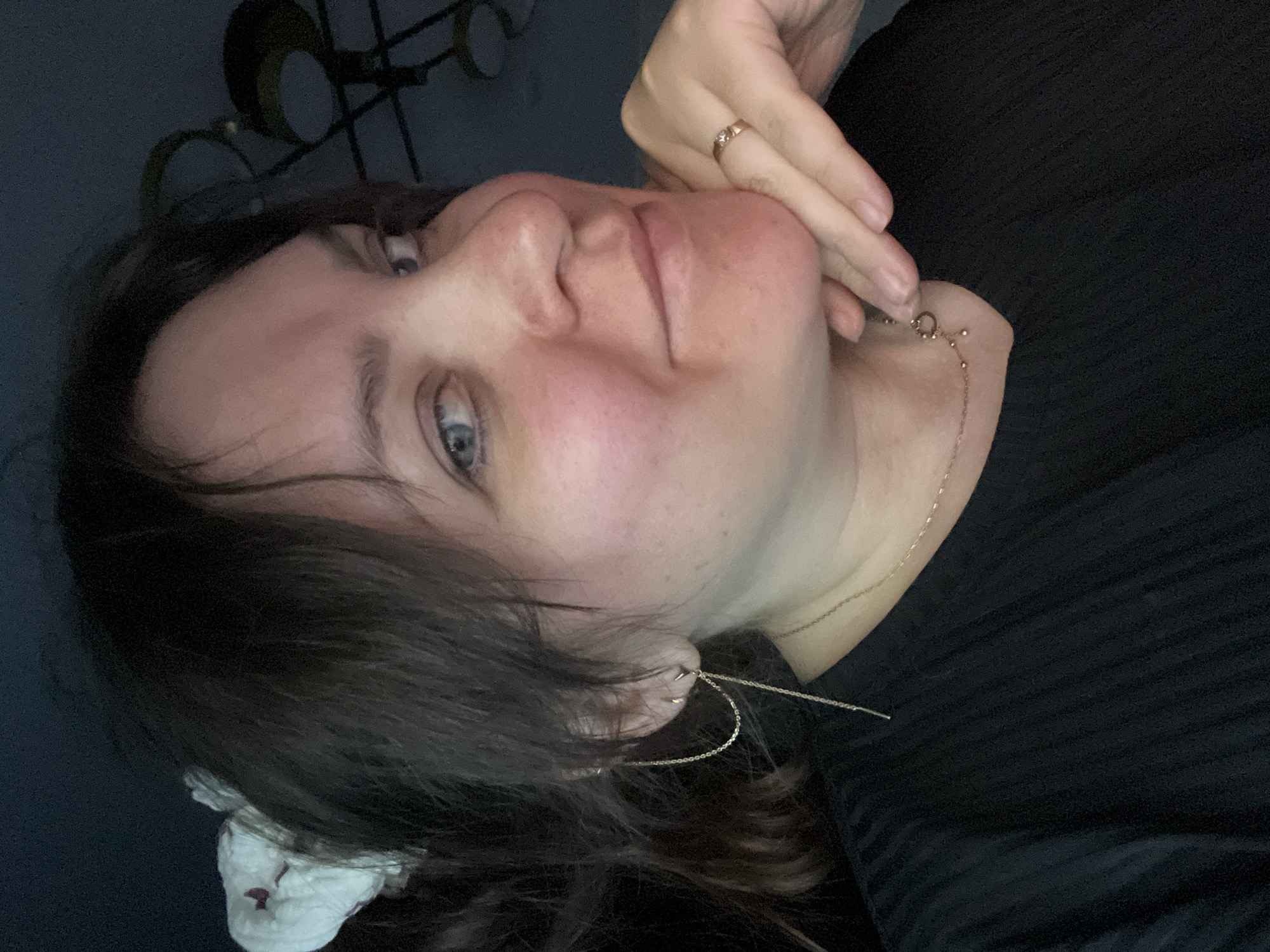 A woman with fair skin and brown hair is pictured in a close-up, low-angle shot. Her hair is partially obscuring her left eye, and she has blue eyes. She is wearing a gold ring on her right hand and a gold necklace. She has an earring that appears to be a double chain. She is wearing a black ribbed shirt. A crumpled white cloth is visible near her hair. The background is dark, with a decorative metal object out of focus. The city of Noviy, Russia is not visible and there is no indication in the photo to suggest it was taken there.