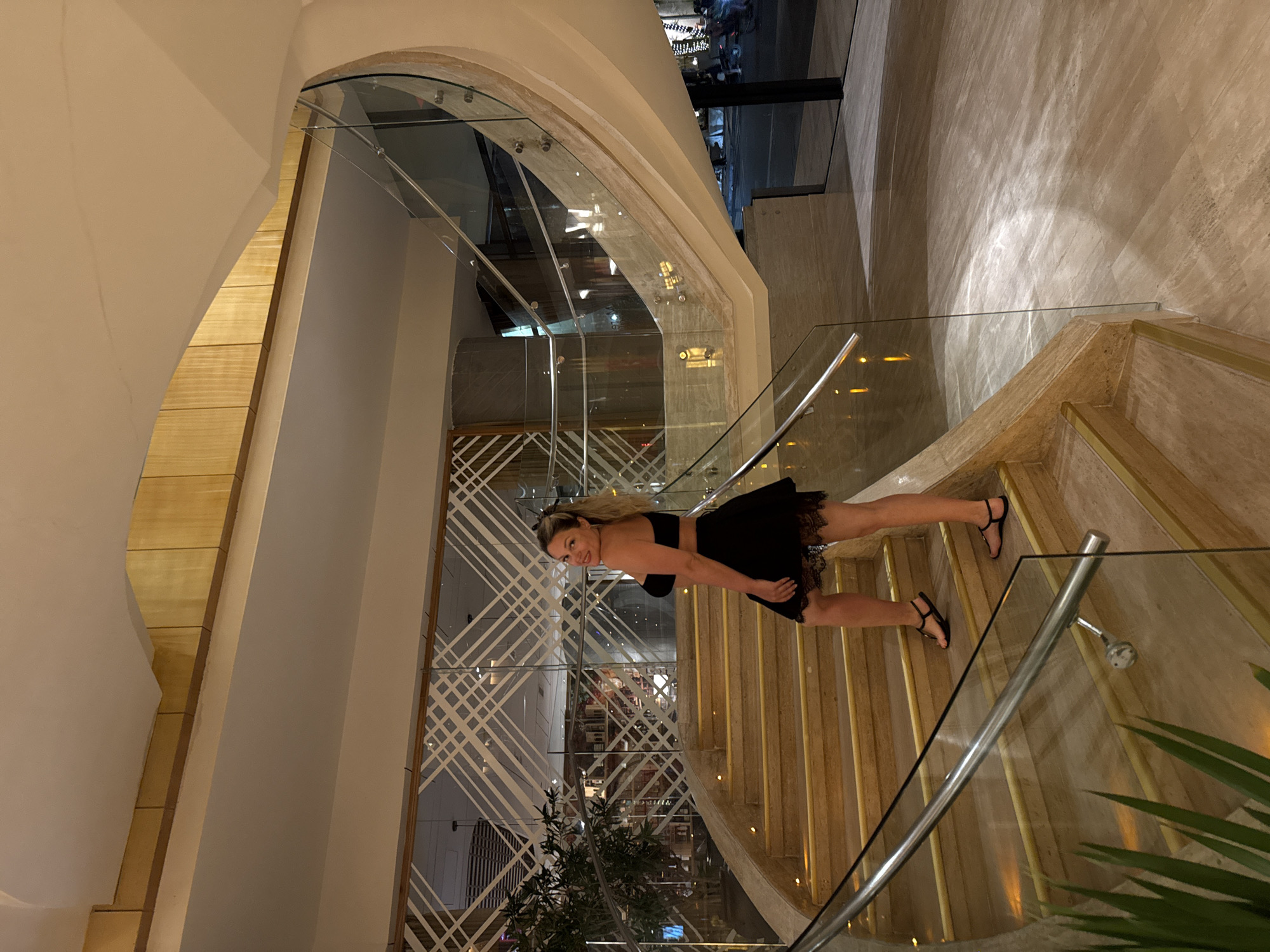A woman stands on marble stairs with glass railings in Nha Trang Ward, Vietnam. She is wearing a black two-piece outfit and sandals. The stairway is inside a building with a patterned glass partition. Outside the building is visible through an arched window.