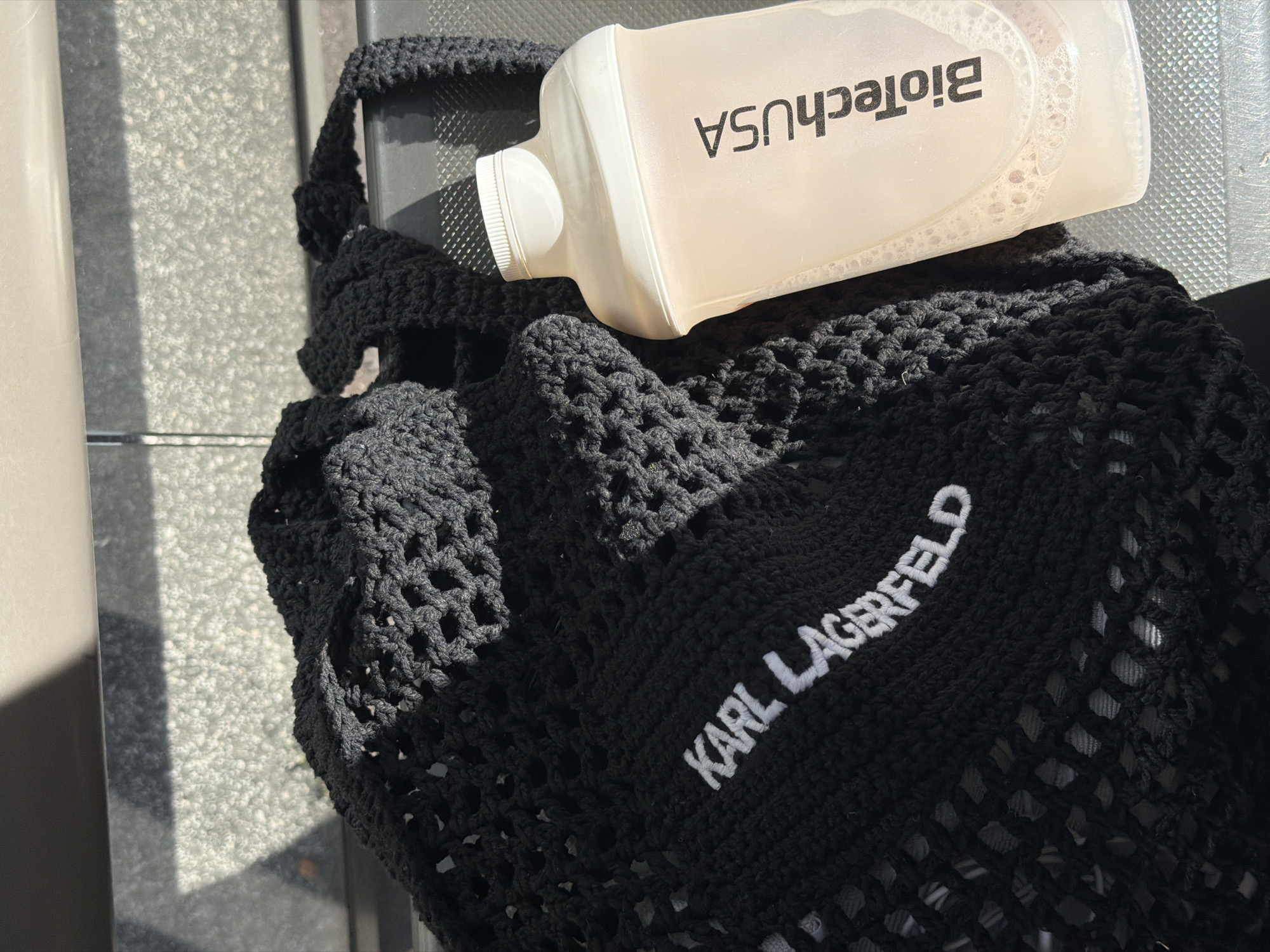 An overhead shot shows a black crocheted bag with "KARL LAGERFELD" embroidered in white on the left side of the bag. To the right of the bag is a pale pink, translucent plastic shaker bottle with a white lid. The bottle has "BioTechUSA" printed vertically in black on its side. Both items are resting on a textured, metallic surface that is partially illuminated by sunlight casting a grid-like shadow. The background is out of focus and appears to be a textured, light-colored surface. There is no indication of the location being Strasbourg, France.