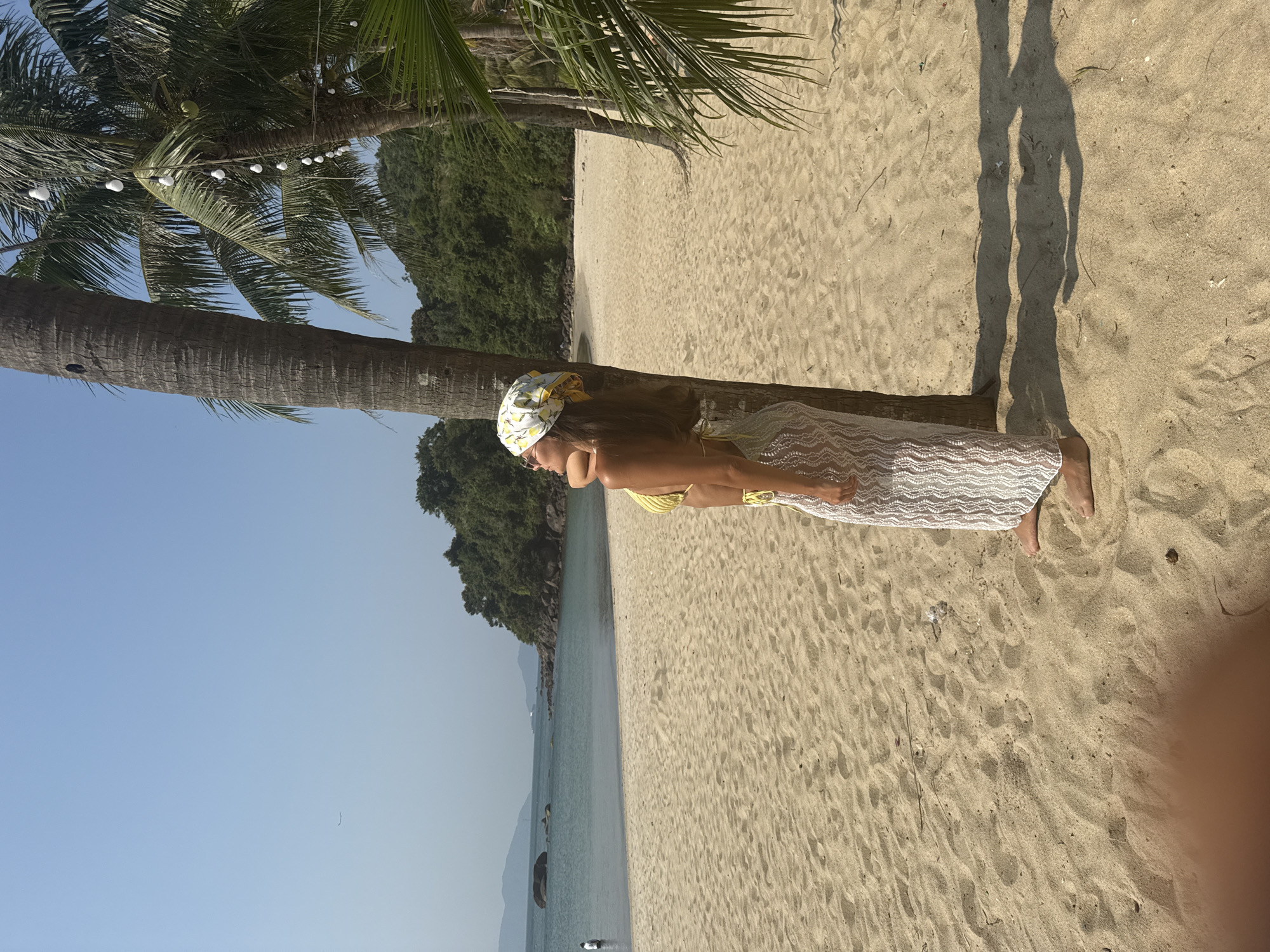 A young woman in a yellow swimsuit and white cover-up leans against a palm tree on a sandy beach. She wears a patterned head covering. The beach is located in Đà Nẵng, Vietnam, as evidenced by the clear blue sky, the tropical foliage behind the beach, and the visible coastline. The water is calm and clear. The scene is brightly lit, and there are shadows on the sand. Some string lights are strung on the palm tree.