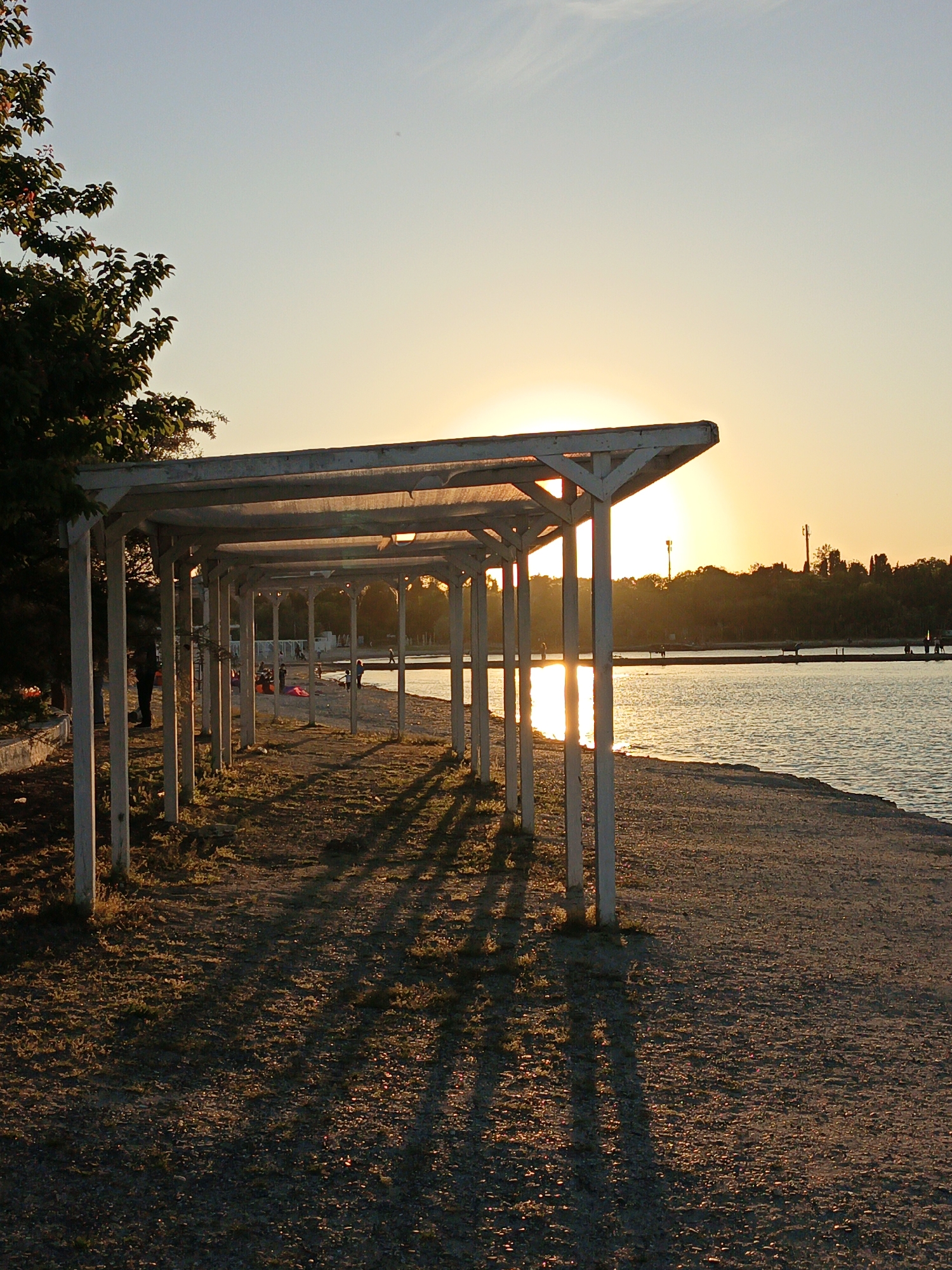 The image depicts a beach scene at sunset in Sevastopol, Ukraine. A long, white wooden pergola structure runs parallel to the shoreline, casting elongated shadows across the sandy and grassy ground beneath. The setting sun is visible through the pergola, reflecting on the water surface, creating a shimmering effect. A few indistinct figures of people can be seen in the background, both on the beach and along a pier or breakwater in the water. A tree is present on the left side of the image. In the distance, there is vegetation, and utility or communication towers can be discerned.