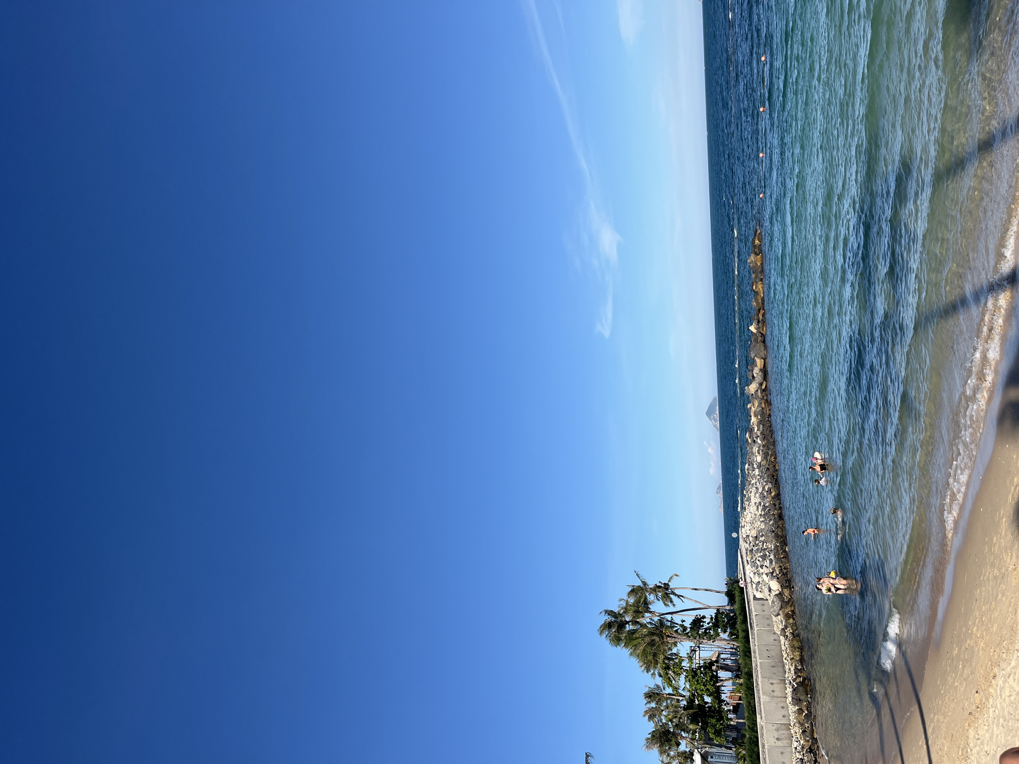 The image depicts a serene beach scene in Bac Nha Trang Ward, Vietnam, under a vast, clear blue sky. The foreground features a sandy beach with gentle waves lapping at the shore. Several individuals are visible in the clear, turquoise water, some swimming and others wading. They appear to be enjoying a leisurely day at the beach.
Along the waterline, a sturdy concrete barrier lined with rocks separates the beach area from a body of water. Beyond this barrier, the sea stretches out to the horizon, dotted with a few small, verdant islands in the distance. Palm trees, characteristic of tropical coastlines, sway gently in the breeze along the shore, adding to the picturesque ambiance. The overall scene conveys a sense of tranquility and natural beauty.