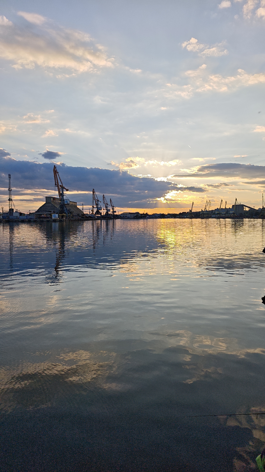 The image captures a sunset view over a harbor in Burgas, Bulgaria. Several industrial cranes line the water's edge on both sides of the frame, silhouetted against the setting sun. A large, pyramid-shaped structure stands prominently among the cranes on the left side of the image. The water reflects the golden light of the sun and the colors of the sky, creating a shimmering effect. The sky is filled with clouds, some tinged with the warm colors of the sunset. The foreground consists of the dark, still water of the harbor.