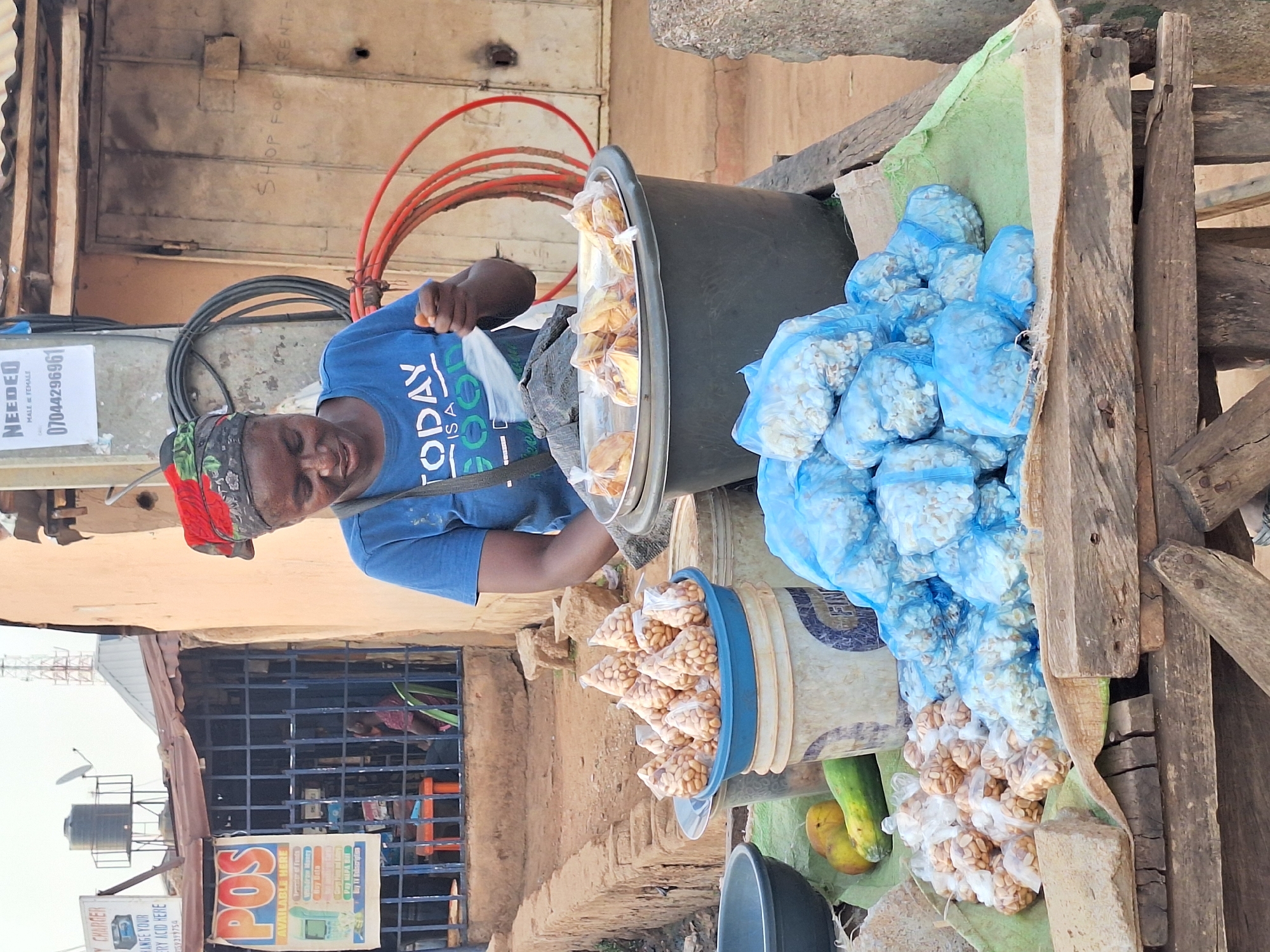 A vendor stands at a wooden table in Sabon Tasha, Nigeria, selling packaged snacks. The vendor is a woman of African descent wearing a blue t-shirt with the word "TODAY" printed on it, and a colorful headscarf. She is looking towards the table, which is covered with a light green cloth. The table holds a large black container filled with what appear to be baked goods in clear plastic bags. Adjacent to the black container are numerous clear blue plastic bags filled with white popcorn. In the foreground is a bucket filled with peanuts in clear plastic bags, and two yellow pears. Behind the vendor, there is a metal barred window and a sign with "POS AVAILABLE HERE" written on it. A faded sign that reads, "NEEDED MALE FEMALE 07044296961" is also visible. A water tank and a telecommunications tower are visible in the background.