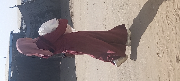A female individual is depicted walking from left to right across an unpaved, light brown sandy surface under clear daylight in Khan Yunis, Palestinian Territory. She is attired in a long-sleeved, full-length dark red or maroon dress cinched with a matching belt, a light pink hijab, and white athletic shoes. A dark shoulder bag is visible on her left side. The individual carries a large, bulky white object, possibly a bundle of fabric or supplies, cradled in both arms. In the background, behind the individual, multiple rudimentary structures constructed from dark tarpaulins and corrugated metal are visible under a bright, clear sky, consistent with an informal settlement or displacement camp environment. A distinct, elongated shadow of the individual is cast on the ground to her right.