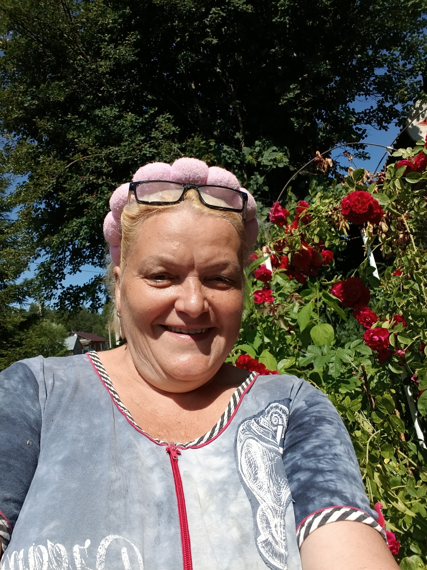 In a medium shot, a fair-skinned woman with blonde hair and light blue eyes smiles and looks directly at the camera. She is wearing a gray zip-up top with a white and black striped trim and a pink head covering adorned with fluffy pom-poms. Black-rimmed glasses rest on her head. Behind her, a vibrant bush of red roses blooms, with lush green foliage and a clear blue sky visible above. A large tree with dark green leaves provides shade in the background. The scene appears to be set in a garden or yard, possibly in the village of Krasnyy Mayak, Russia, given the presence of a small, rustic house visible through the trees on the left. The sunlight suggests a warm, summer day.