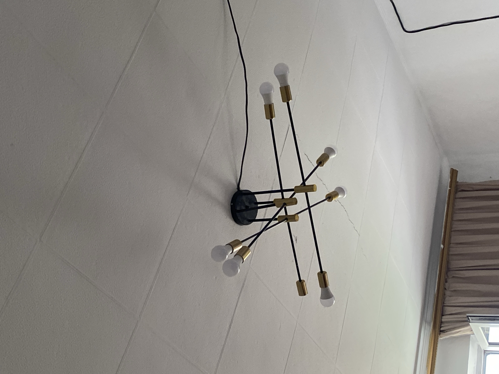 The image shows a black and gold sputnik chandelier with eight white LED bulbs hanging from a textured white ceiling. The chandelier has multiple arms extending outwards from a central black base. The ceiling appears to be made of acoustic tiles and shows some minor cracking. A black electrical cord runs from the chandelier across the ceiling. In the lower portion of the image, a beige curtain is visible, partially obscuring a window. The location is specified as Qaraoy, Kazakhstan.