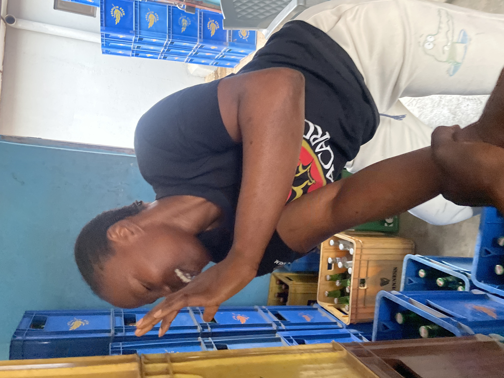 A person wearing a black t-shirt and white pants is leaning forward among several stacks of beer crates. The crates are primarily blue and yellow, some containing beer bottles. The background includes a blue wall and stacks of blue crates. The location is Jos, Nigeria.