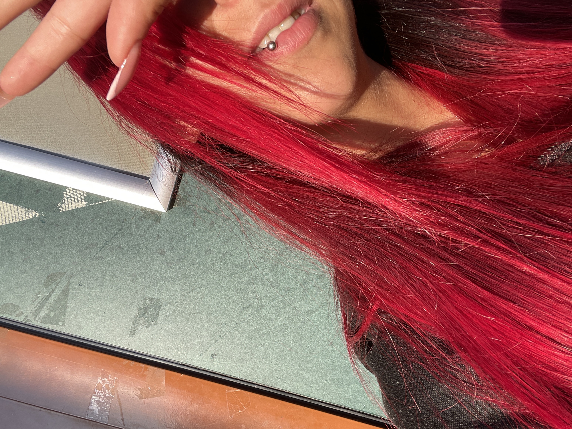 A close-up shot shows a person with vibrant red hair, partially obscuring their face. A hand with manicured nails is held up, shielding their eyes from the sun. A lip piercing with a silver stud is visible on their lower lip. The background consists of two distinct surfaces: a reddish-brown textured area on the left and a muted green textured surface on the right, possibly an outdoor urban environment. The lighting suggests it is daytime, with strong sunlight creating shadows. There is no information in the image to suggest this location is in Sofia, Bulgaria.